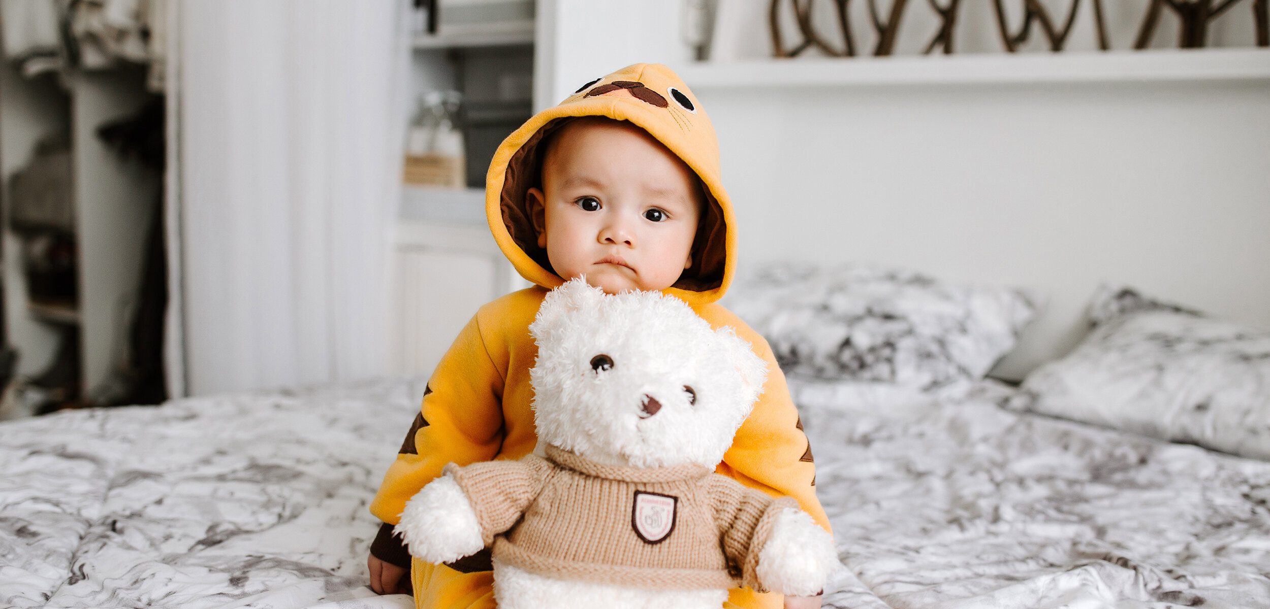 Gorgeous baby in soft yellow clothes hugging teddy bear on bed