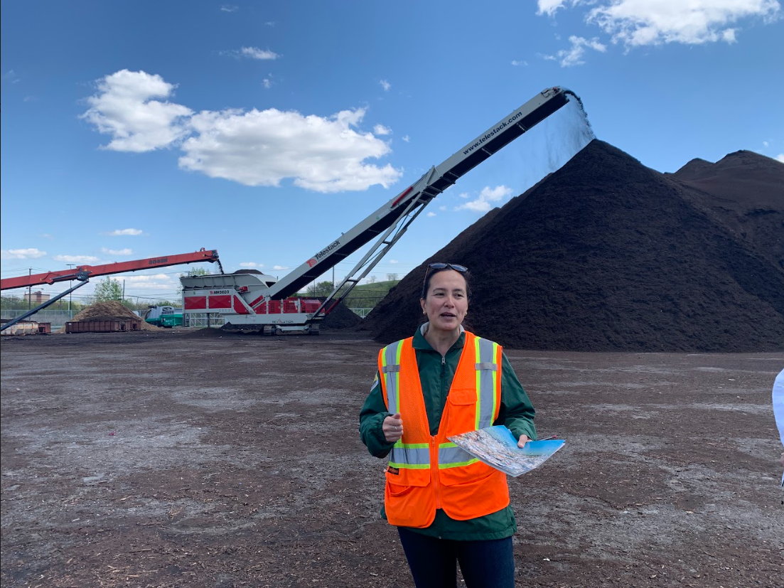   Marguerite stands before a conveyor of sifted NYC yard waste and food scraps.  In 2022 $19 million was budgeted to support curbside collection and food scrap drop-offs (FSDOs), $7.5 million for collection in schools, and $4 million for fall, leaf a