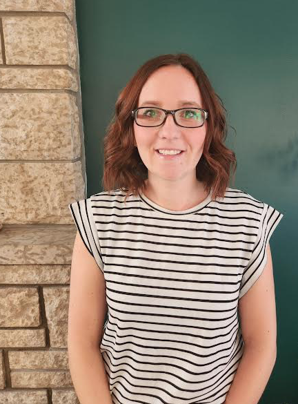 Young woman with shoulder-length wavy brown hair, wearing glasses and a white and black striped short-sleeve top, smiling indoors against a brick wall and teal background.