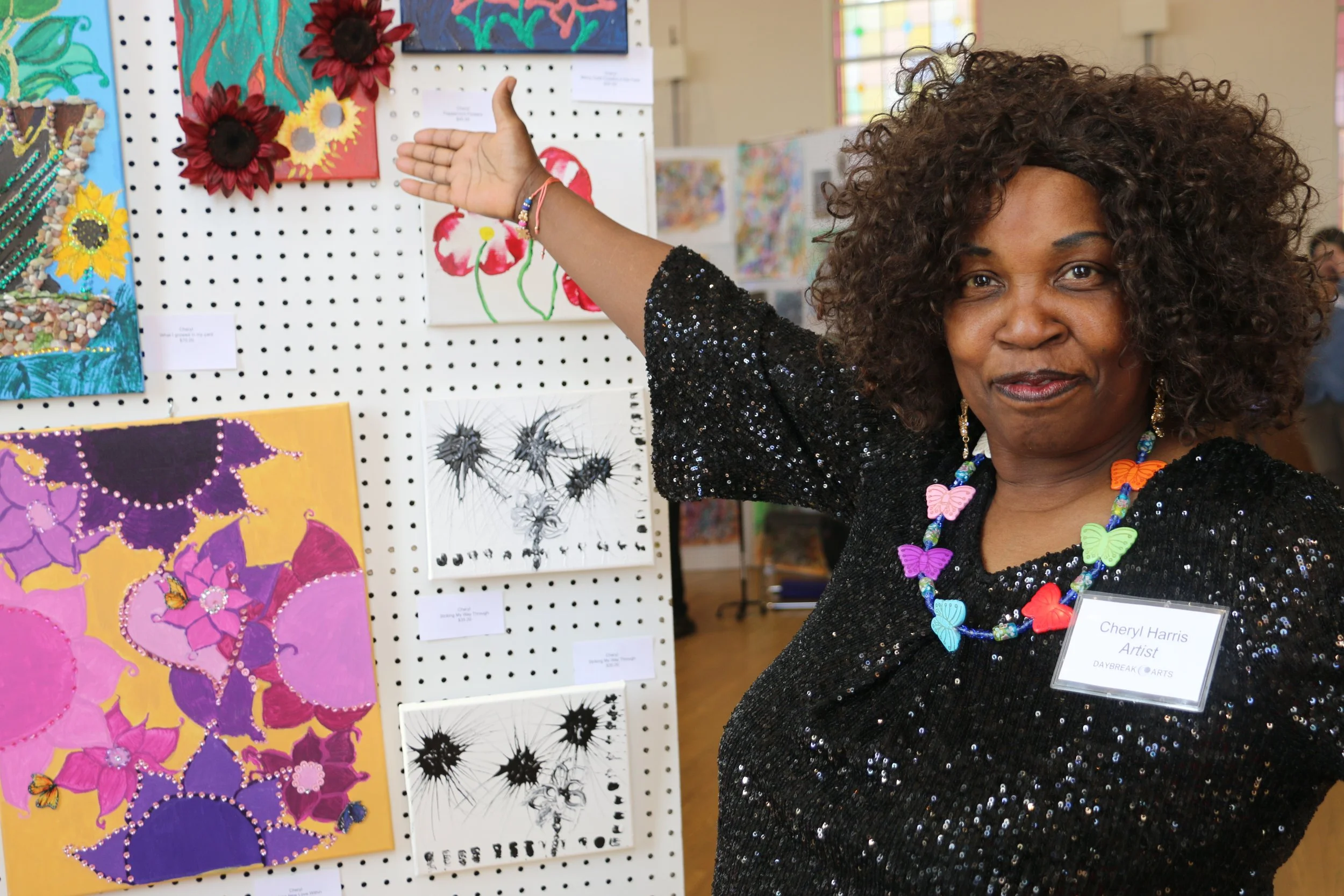 Woman with curly hair and butterfly necklace at art exhibit, pointing to colorful artwork on pegboard display.