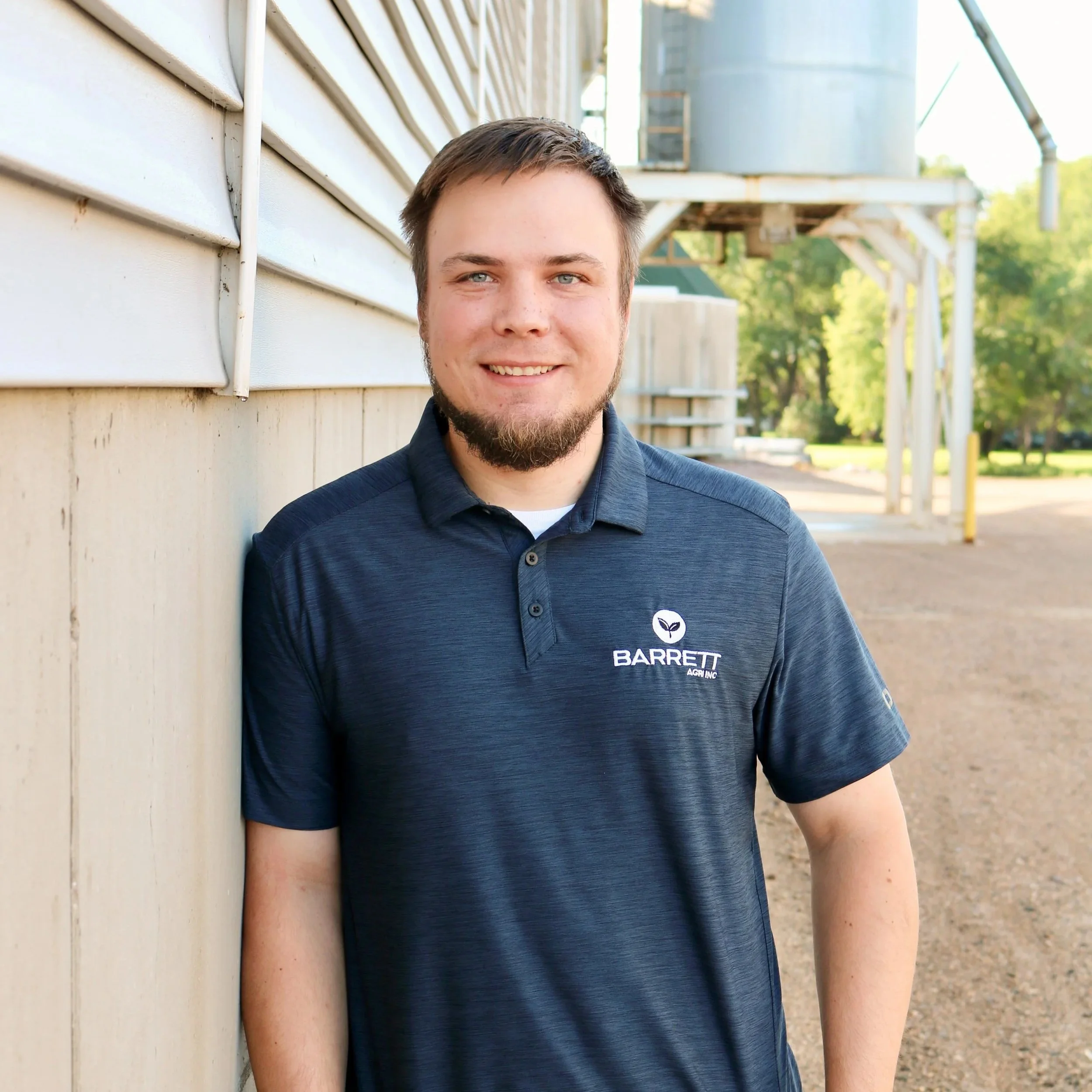 A young man with light skin, short brown hair, and a beard, smiling and leaning against a wall outside. He is wearing a navy blue polo shirt with a logo that says 'Barrett Agri Inc.'