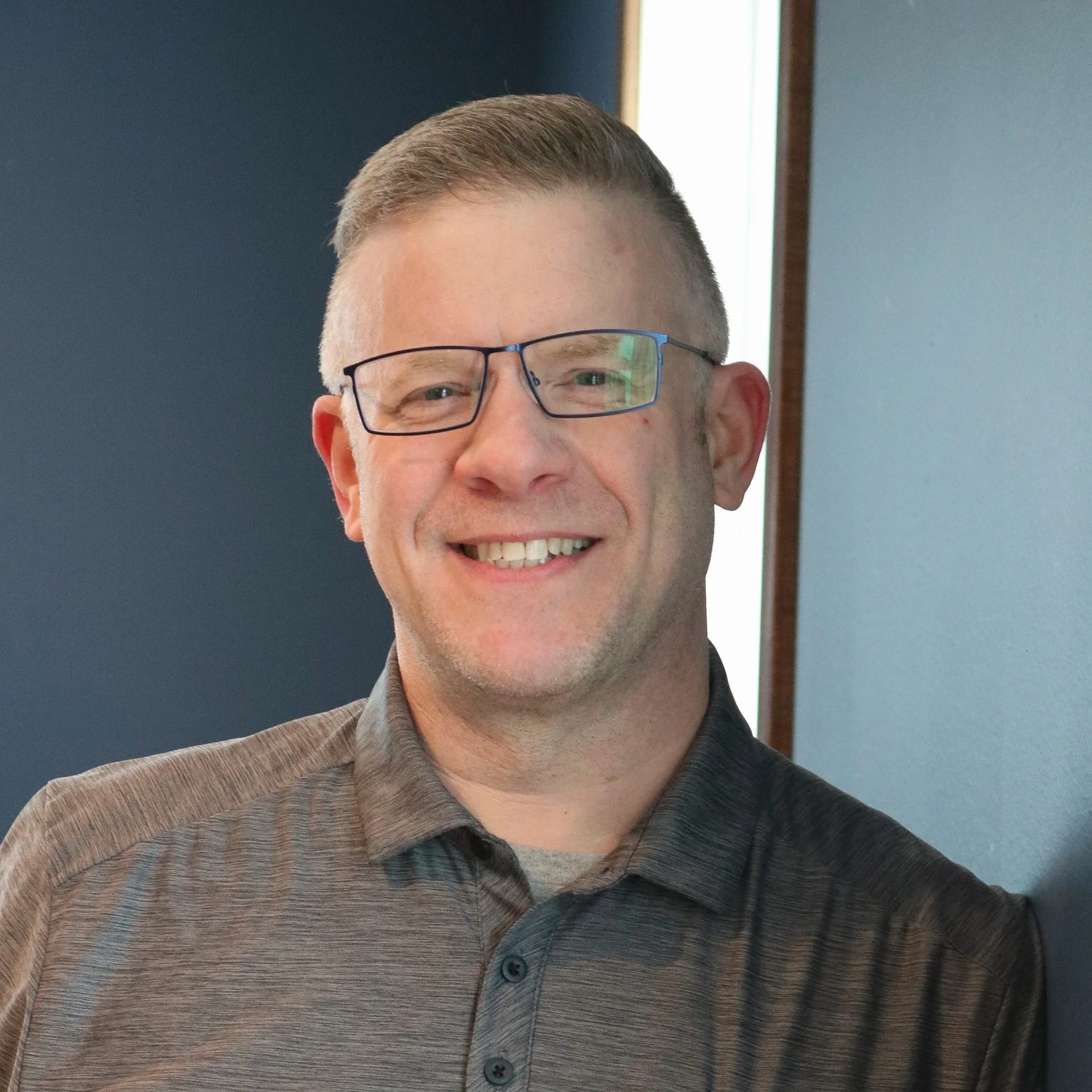 A smiling man with glasses wearing a gray striped collared shirt, standing indoors against a blue and light-colored wall.