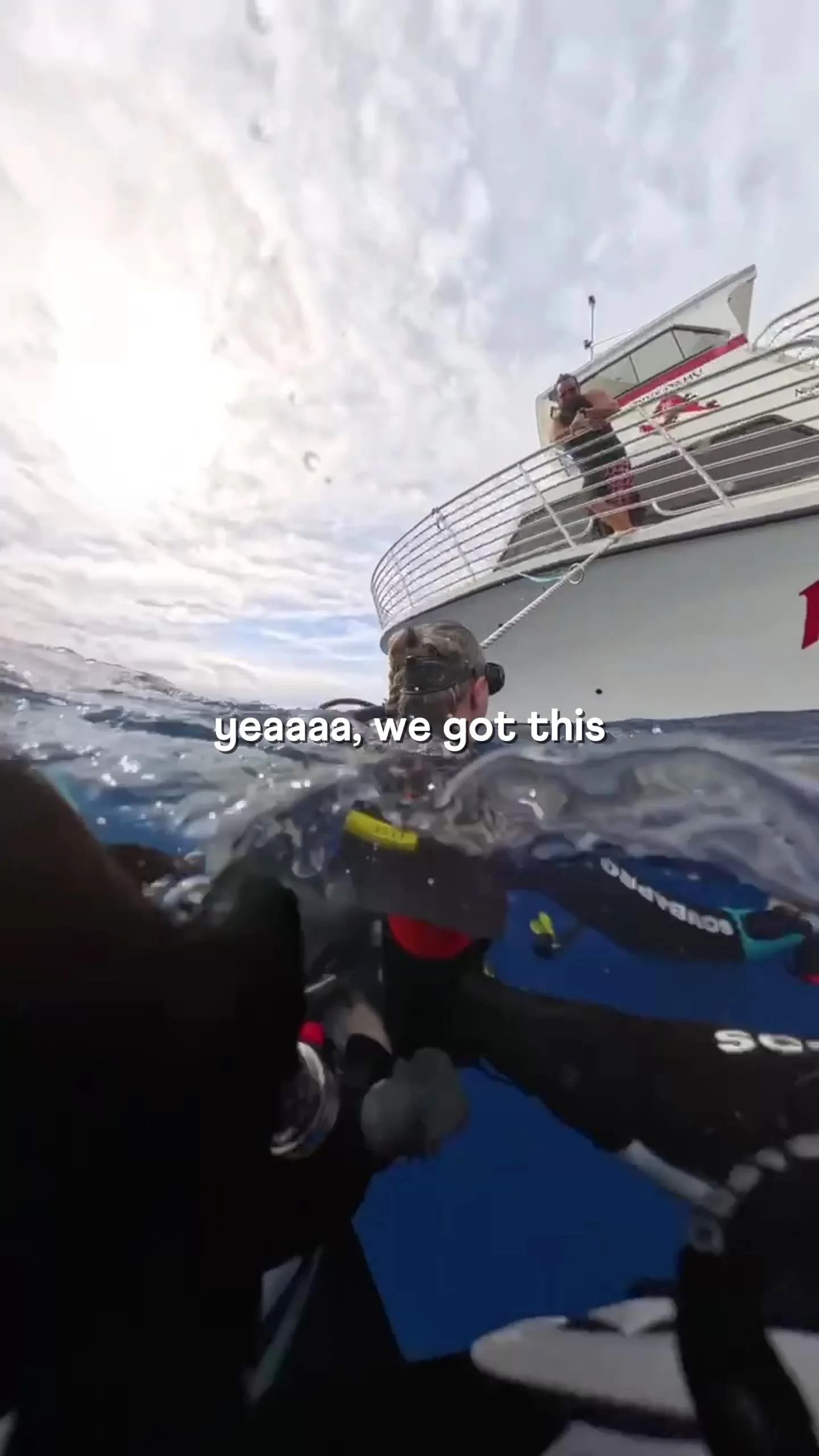 A person in wetsuit and snorkeling gear in the water holding onto a boat ladder, with a person on the boat watching and the sky cloudy above.