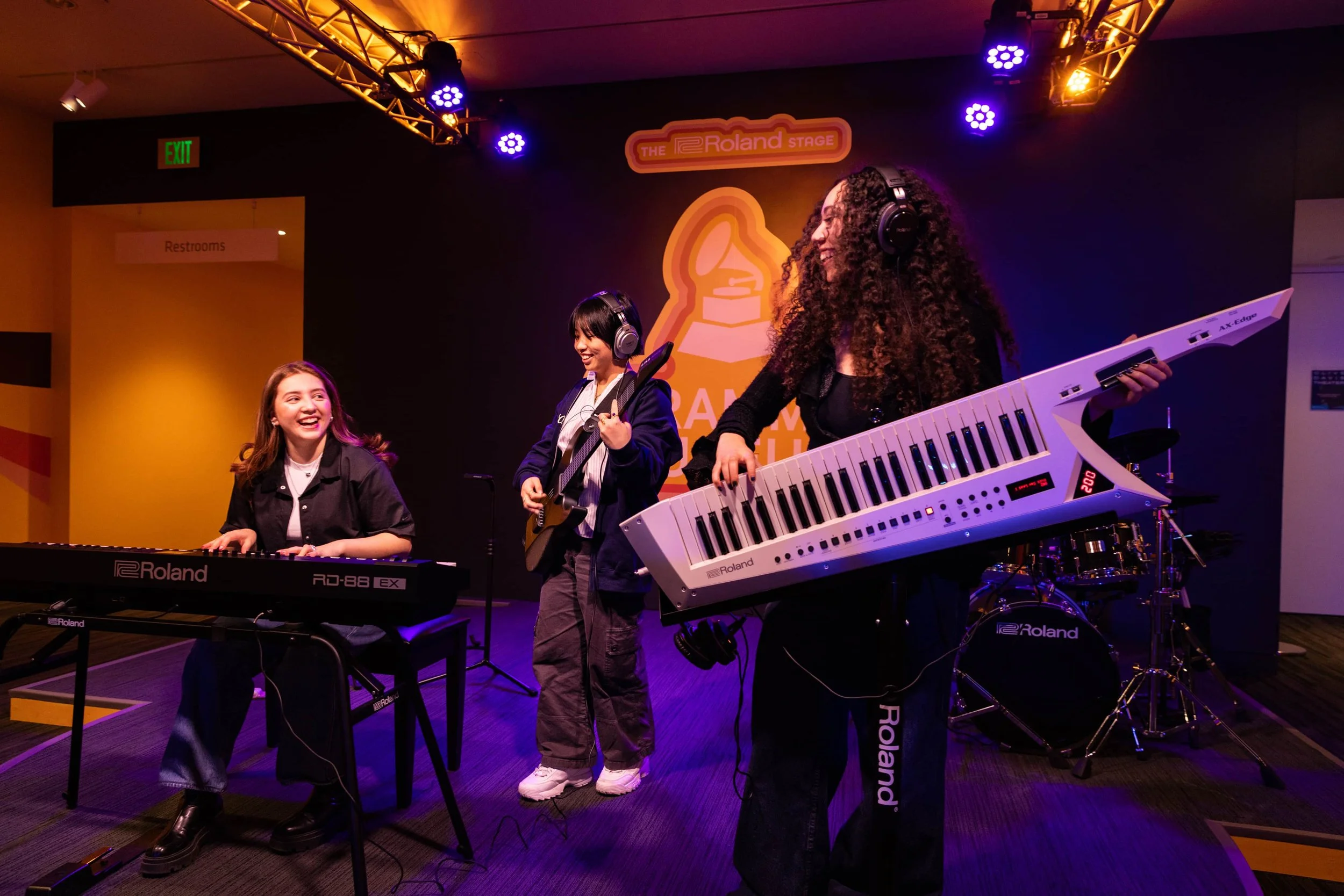 Three women perform music on stage with keyboards and guitar, under colorful stage lighting, with a backdrop displaying a logo and the words 'The Roland Stage'.