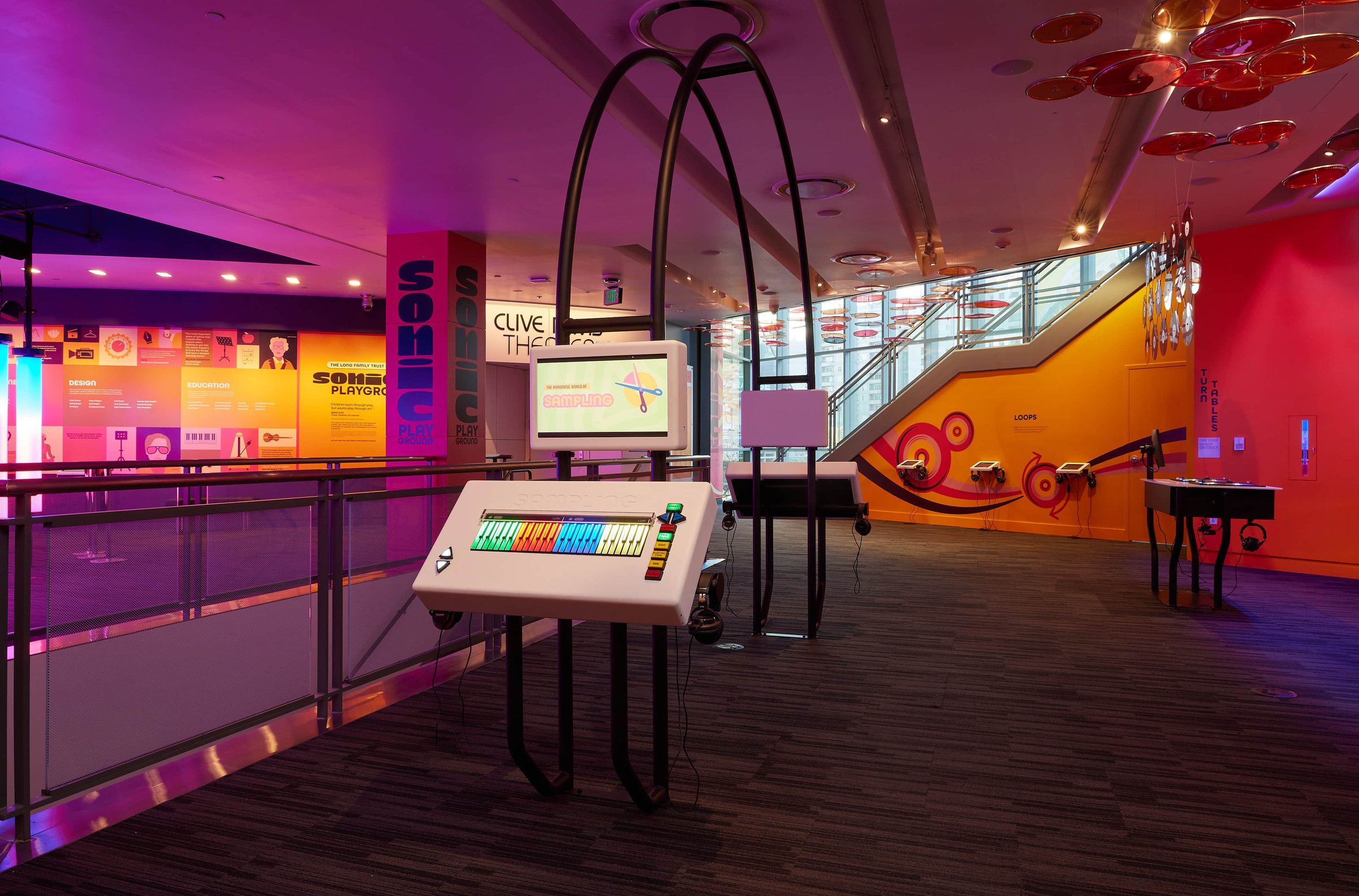 Interior of a colorful interactive science or technology exhibit with displays, monitors, and equipment on the floor and walls, in a modern building with large windows and staircase.