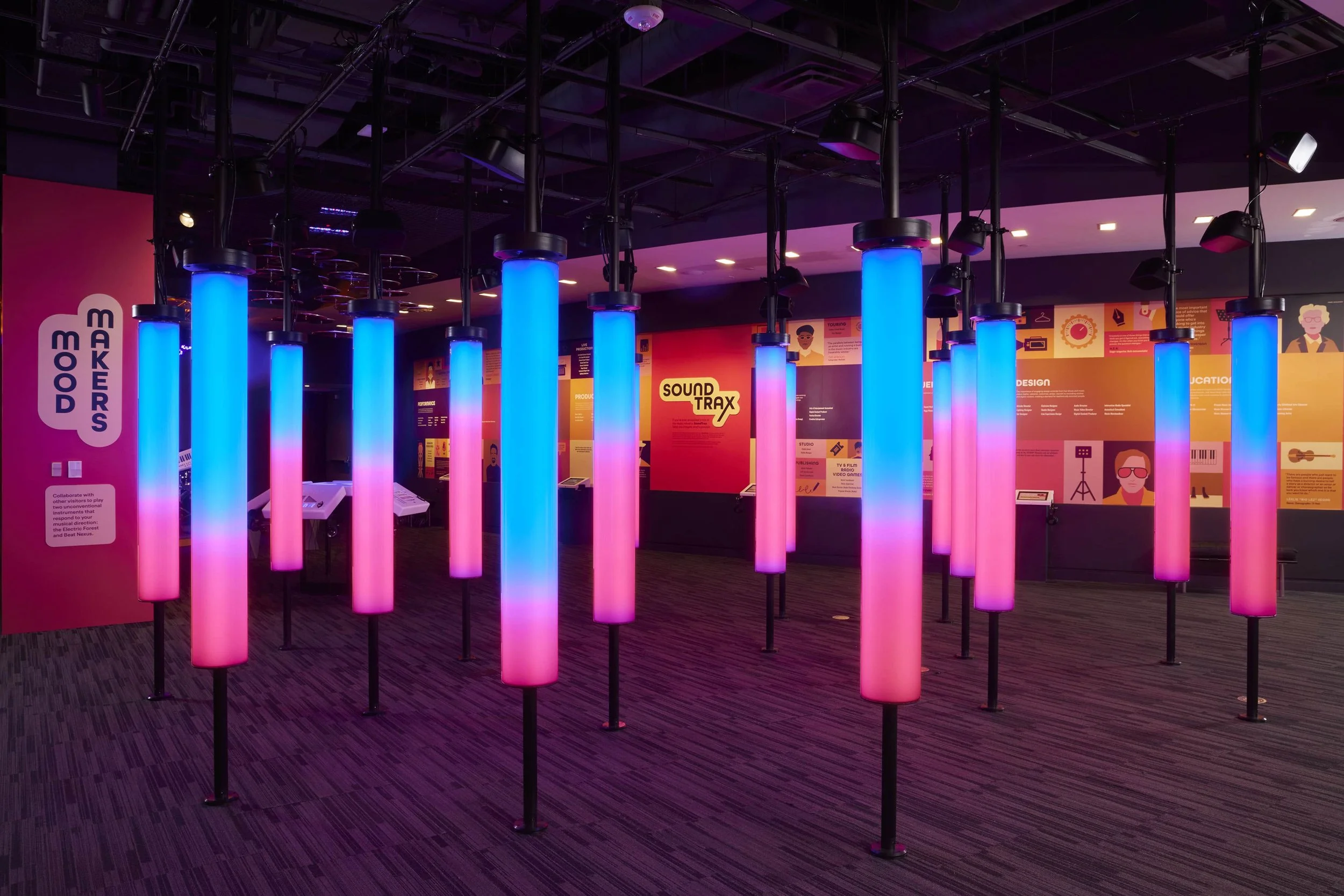An exhibit with vertical LED light tubes emitting pink and blue lights, arranged in a grid pattern. The background has colorful posters and a sign that reads 'SOUND TRAX'.
