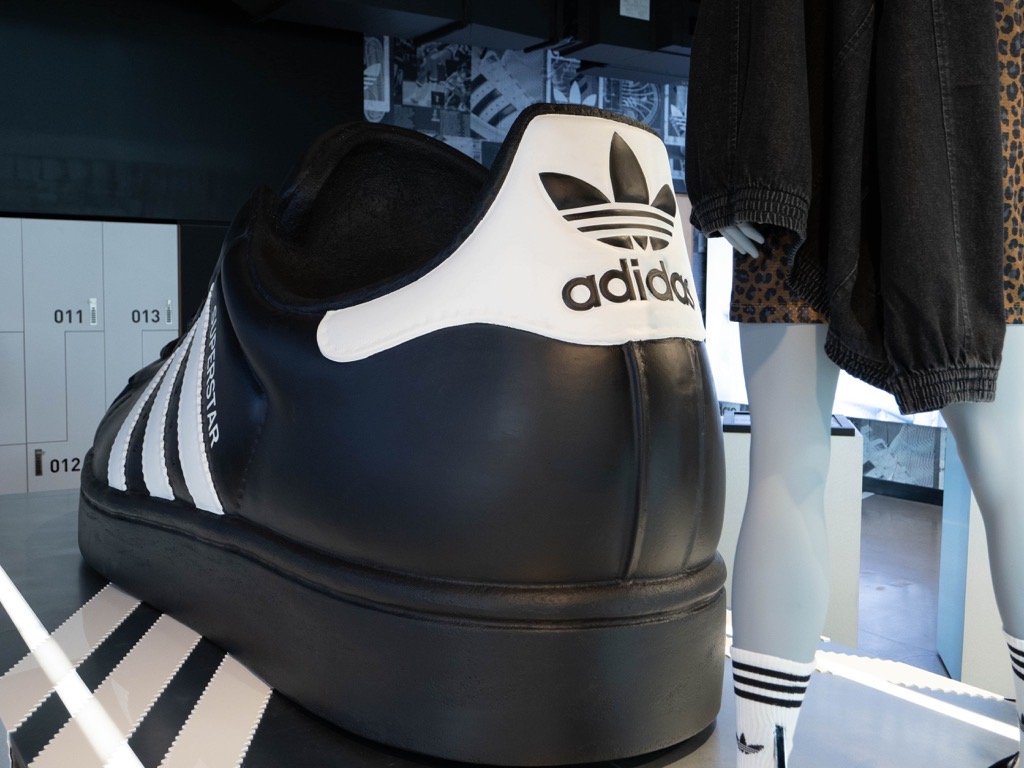 Adidas Originals Big Logo sneaker displayed on a platform in a retail store alongside mannequin legs dressed in black shorts, striped socks, and sneakers, with lockers and patterned decor in the background.