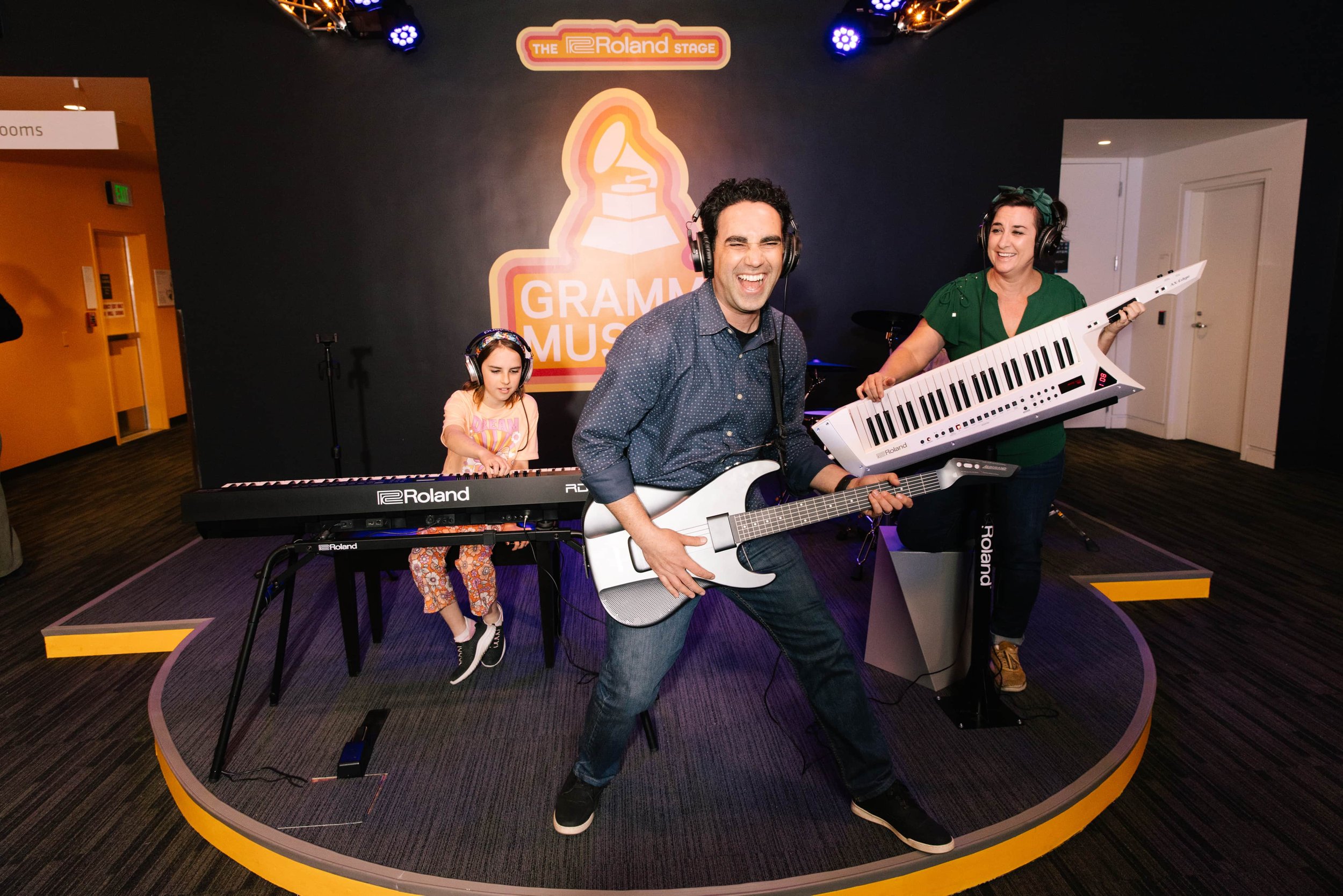 A group of three musicians performing on a stage at the Grammy Museum, with a logo sign behind them. The man in the middle is playing an electric guitar and singing, the girl on the left is playing a keyboard, and the woman on the right is playing a 