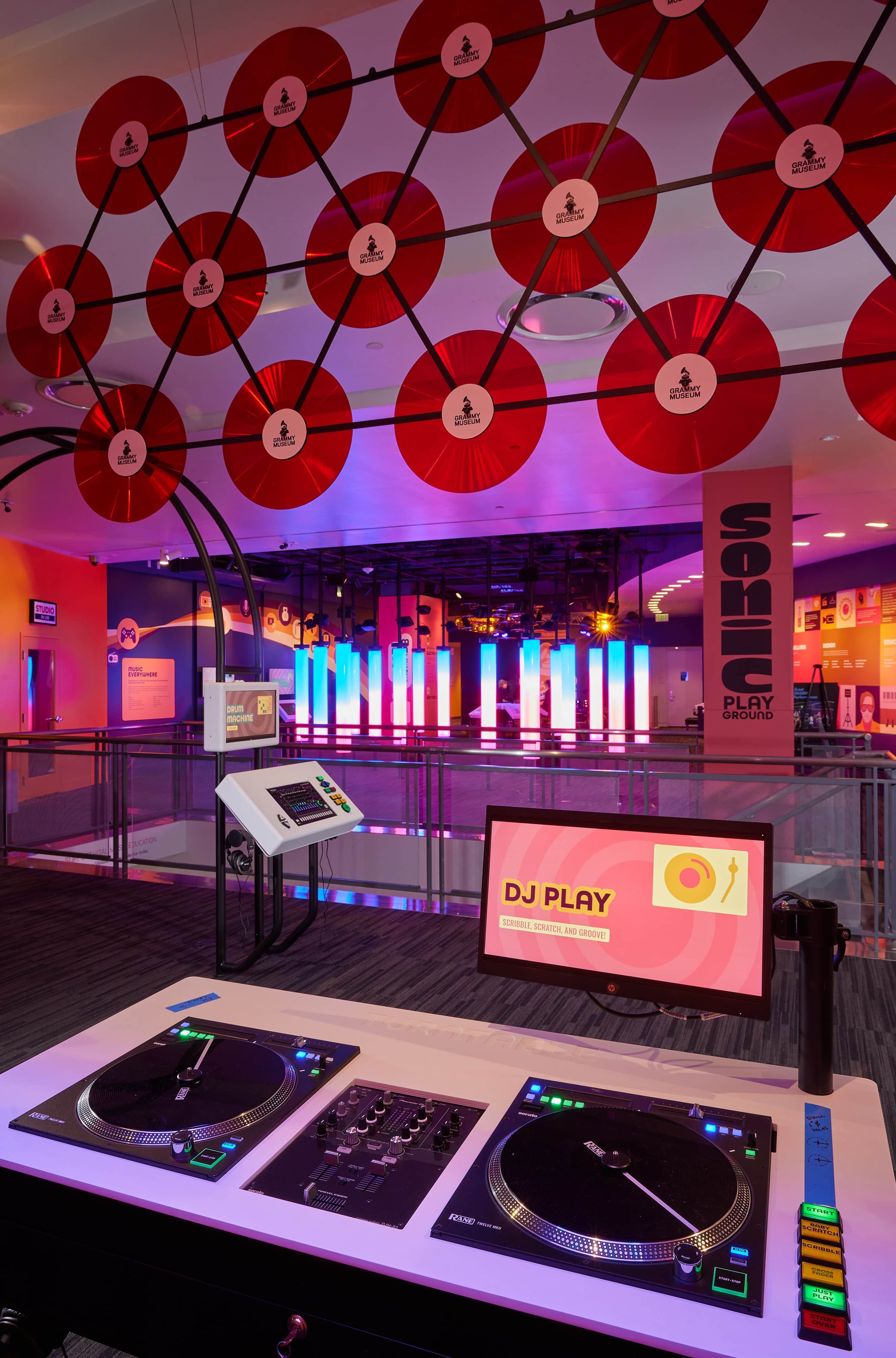 A DJ setup with turntables and a mixer, a monitor screen that reads 'DJ Play' with a record icon, in a vibrant indoor entertainment space with colorful lighting, hanging red discs, and a sign indicating a splash play area.