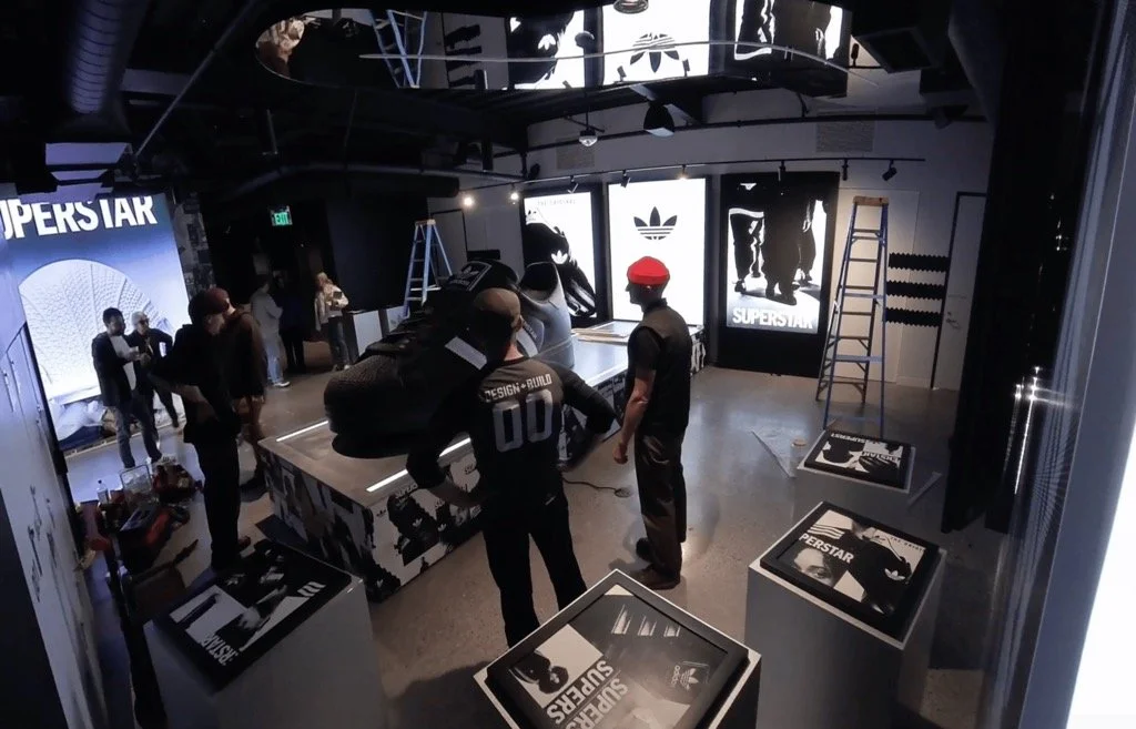 People assembling and setting up an Adidas SuperStar sneaker display in a retail store or showroom, with large illuminated Adidas logos and posters in the background.