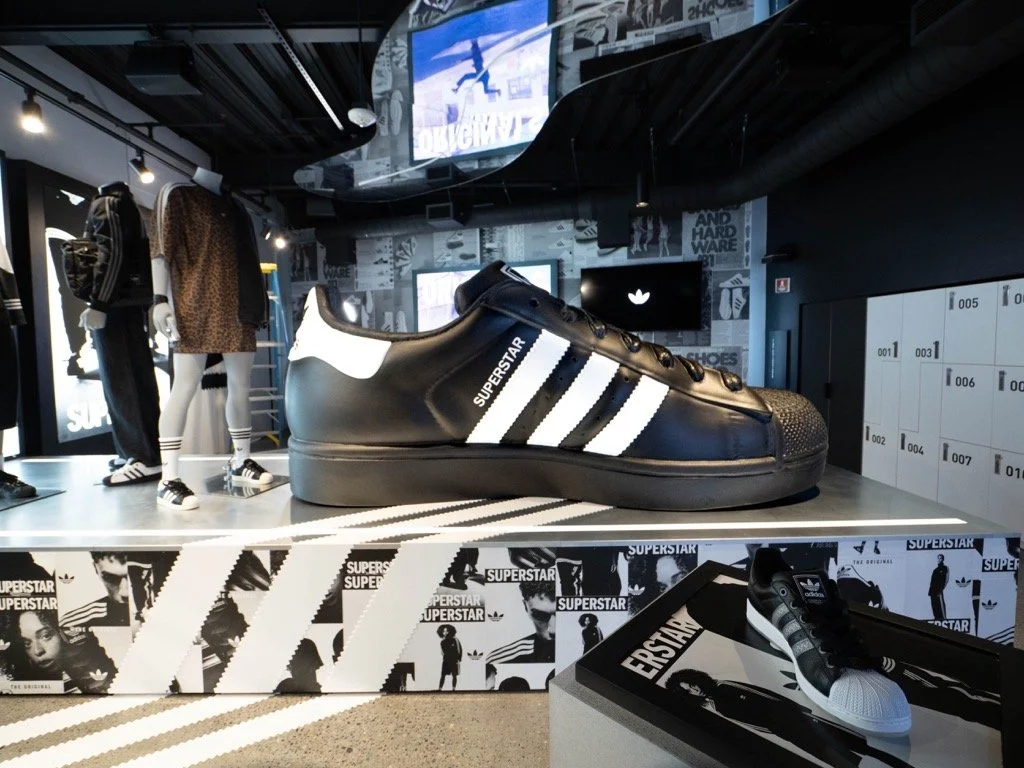 Large black Adidas sneaker with white stripes and the word 'Superstar' on the side, displayed in a retail store with mannequins dressed in athletic clothing.
