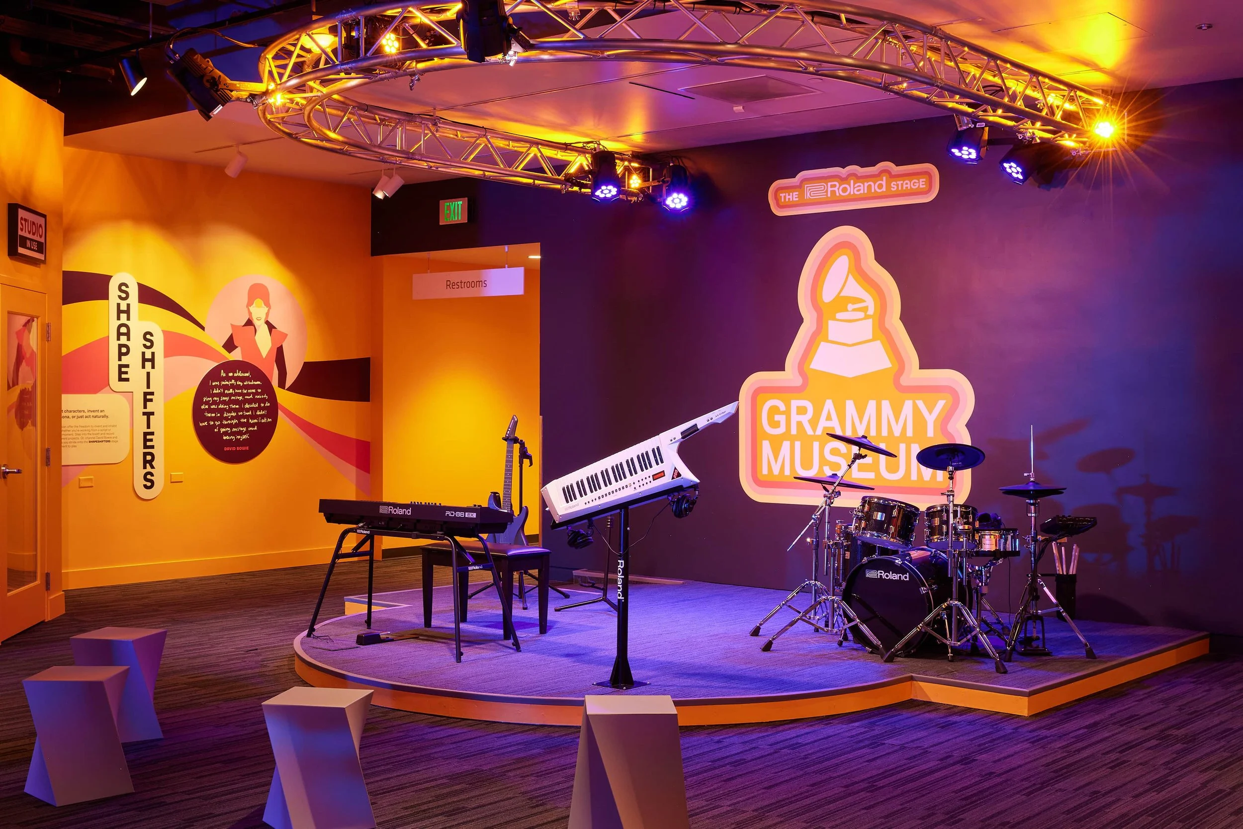 Stage at the Grammy Museum with musical instruments including a keyboard, drum set, and a microphone. The background features a large sign with a gramophone icon and the words "GRAMMY MUSEUM" illuminated, with purple and yellow lighting effects.
