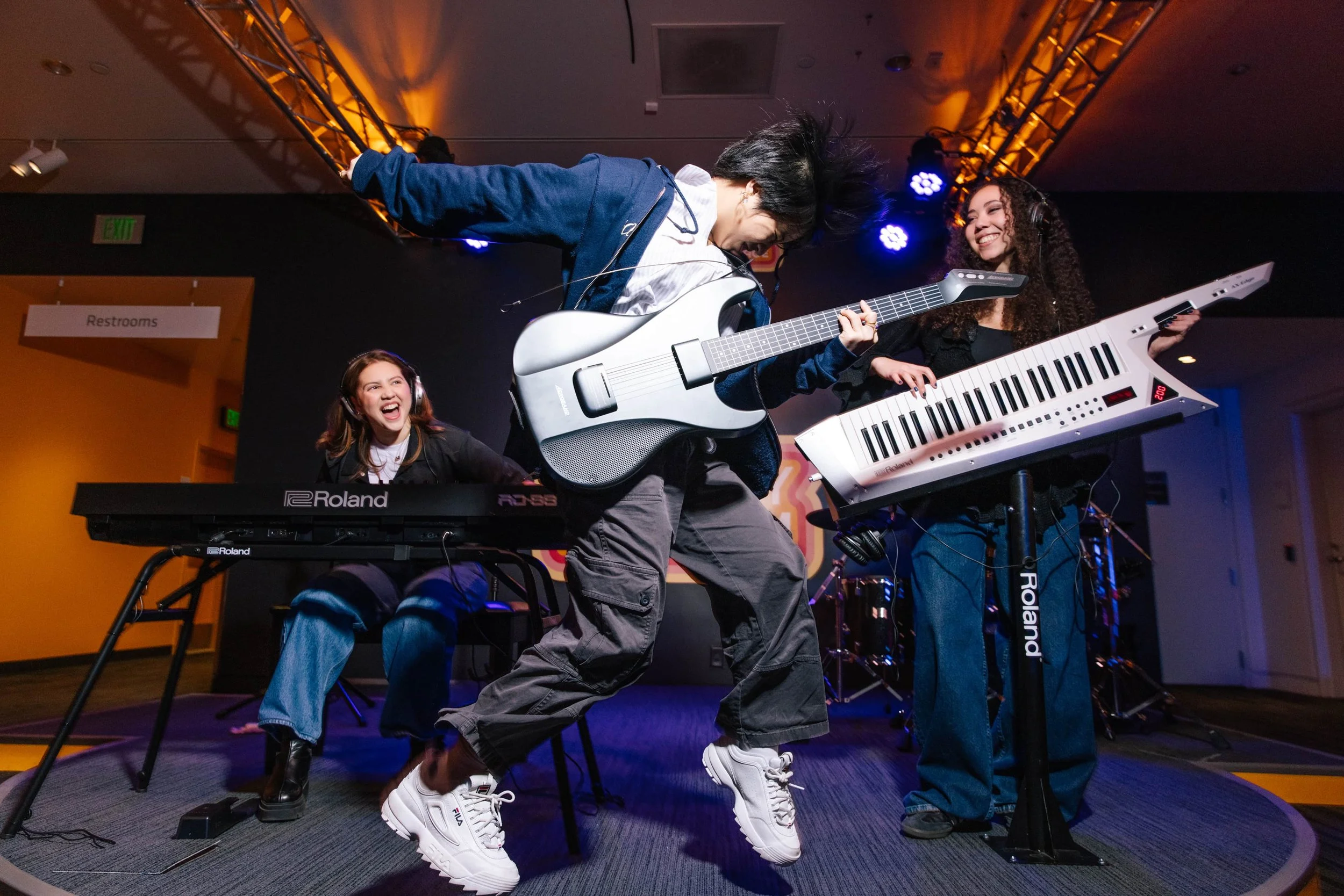 Three young women playing musical instruments on stage: one with a keyboard, one with an electric guitar, and one with a keytar, all smiling and having fun.