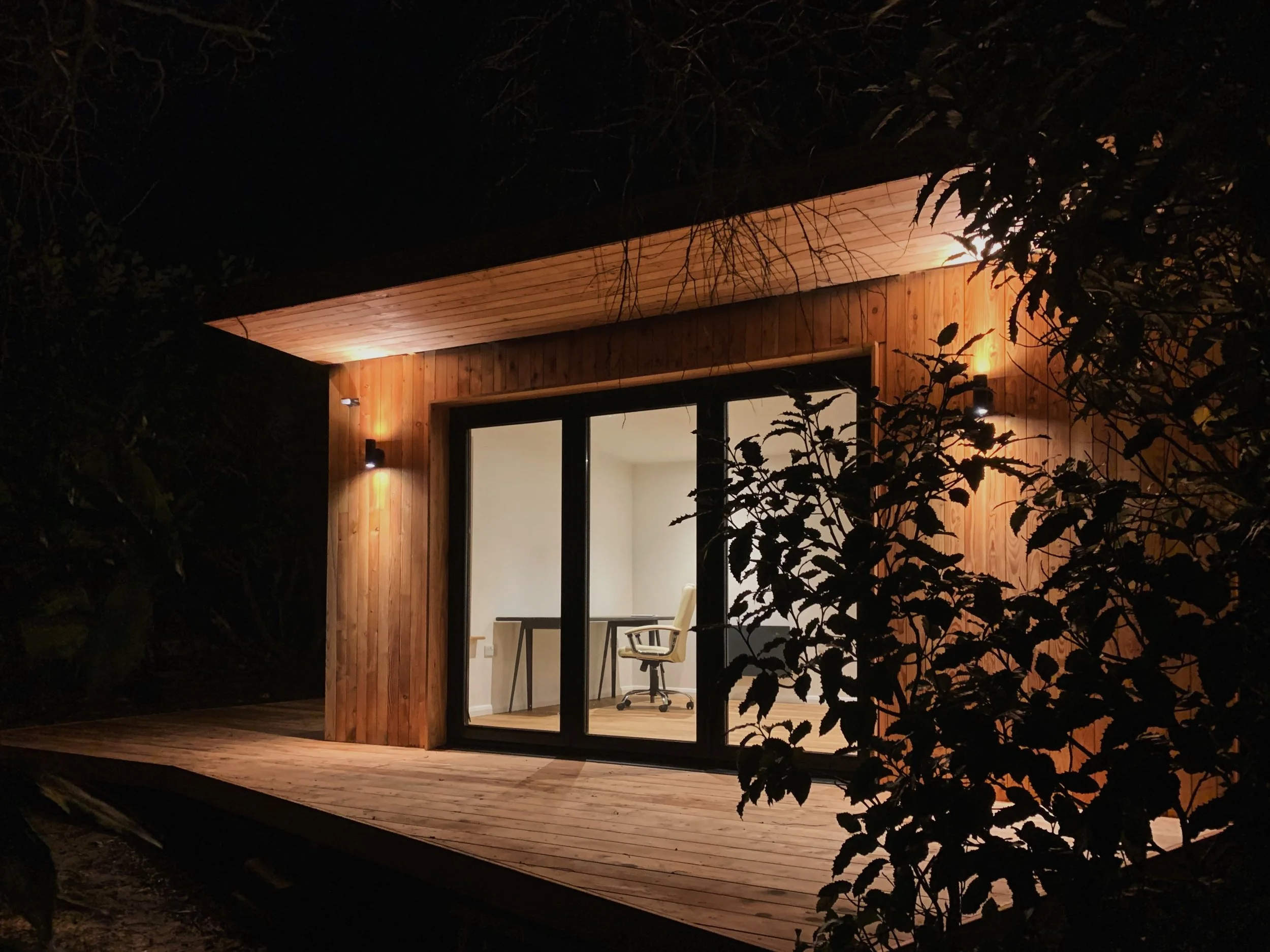 A modern tiny house with a wooden exterior, illuminated from the inside, with large glass sliding doors showing a white desk and an office chair inside. The house sits on a wooden deck surrounded by dark foliage.