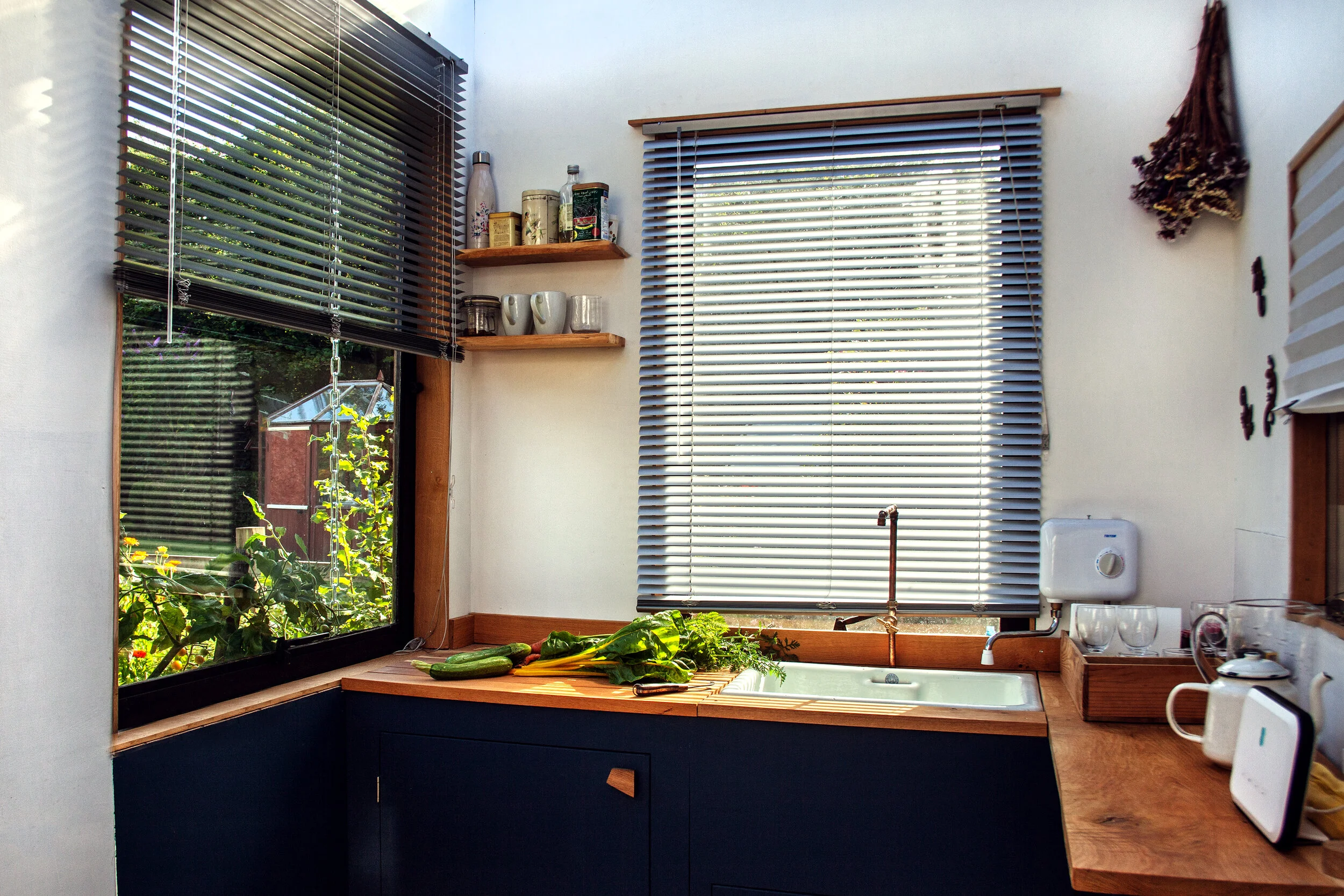 Kitchen corner with large window, window sills, and wooden countertop, fresh vegetables on the counter, hanging herbs, and kitchen utensils.