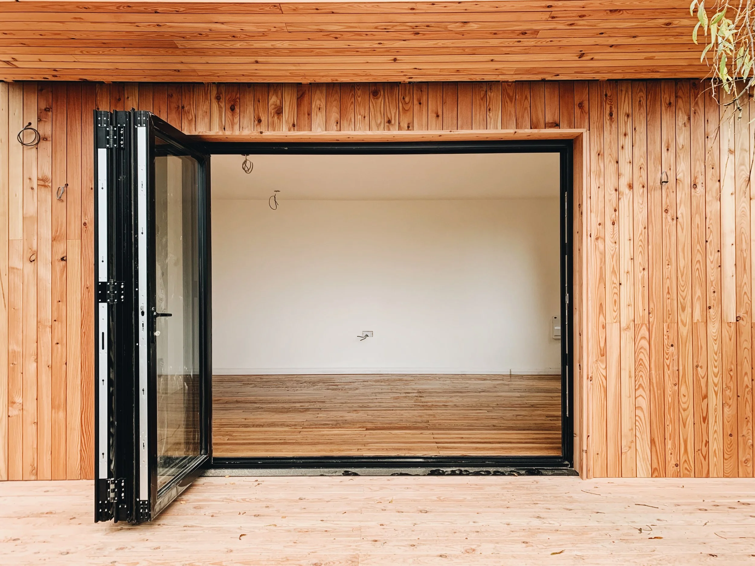Interior of a room with wooden walls and flooring, an open black-folding glass door, and a white wall with electrical wiring visible.