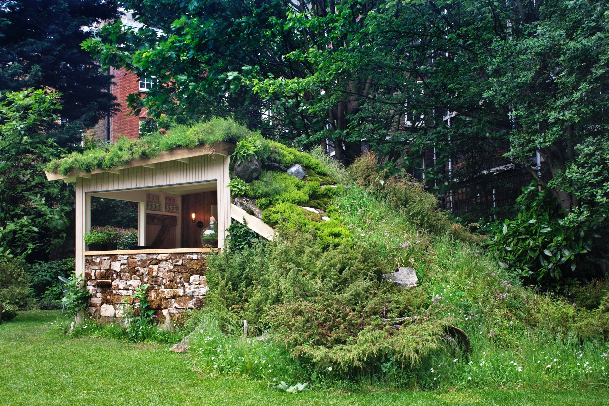 Small house built into a grassy hillside with a sloped roof covered in grass and plants, surrounded by lush green trees and bushes.