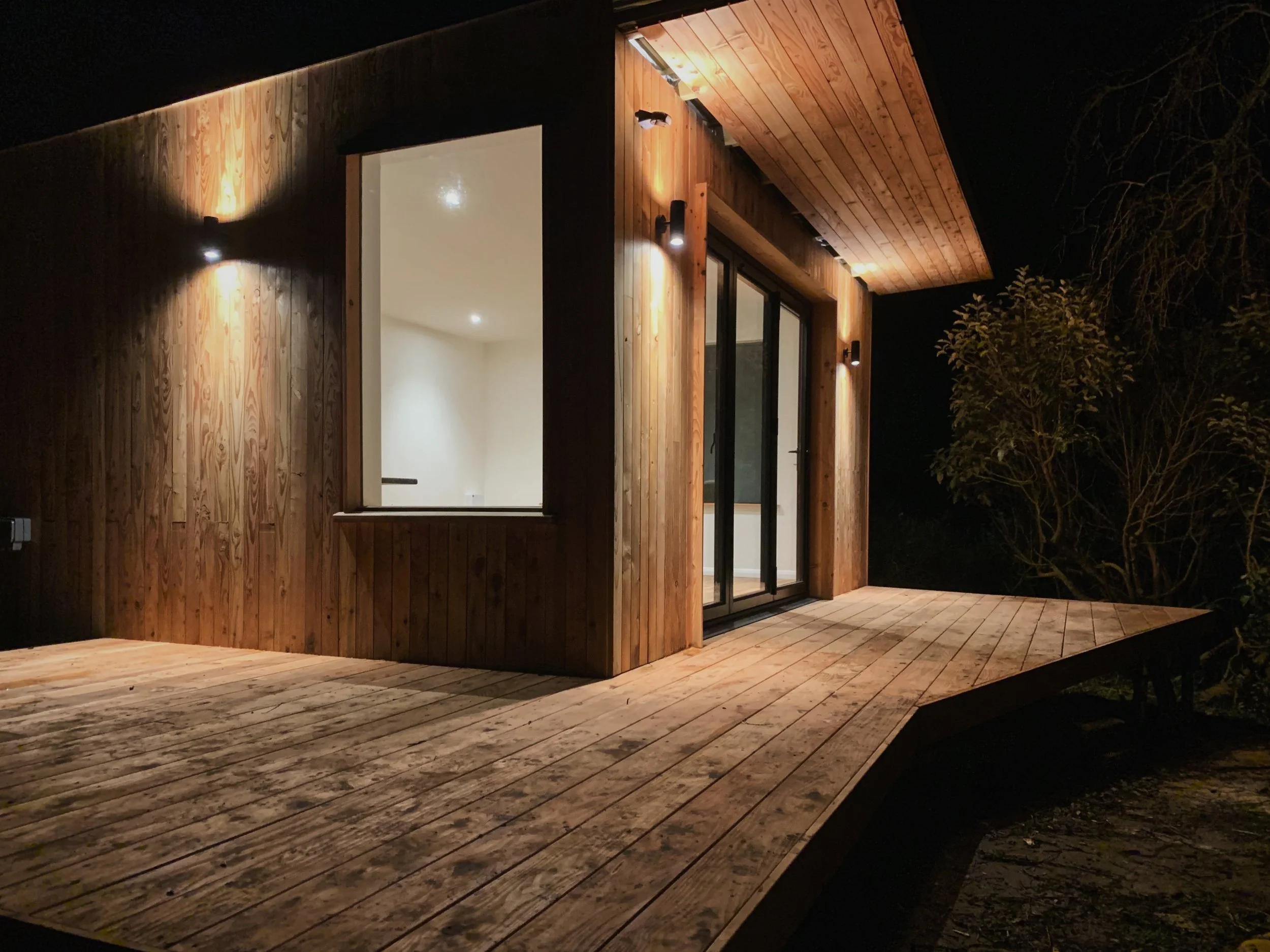 A modern wooden house with a large sliding glass door, exterior lighting, and a wooden deck during nighttime.