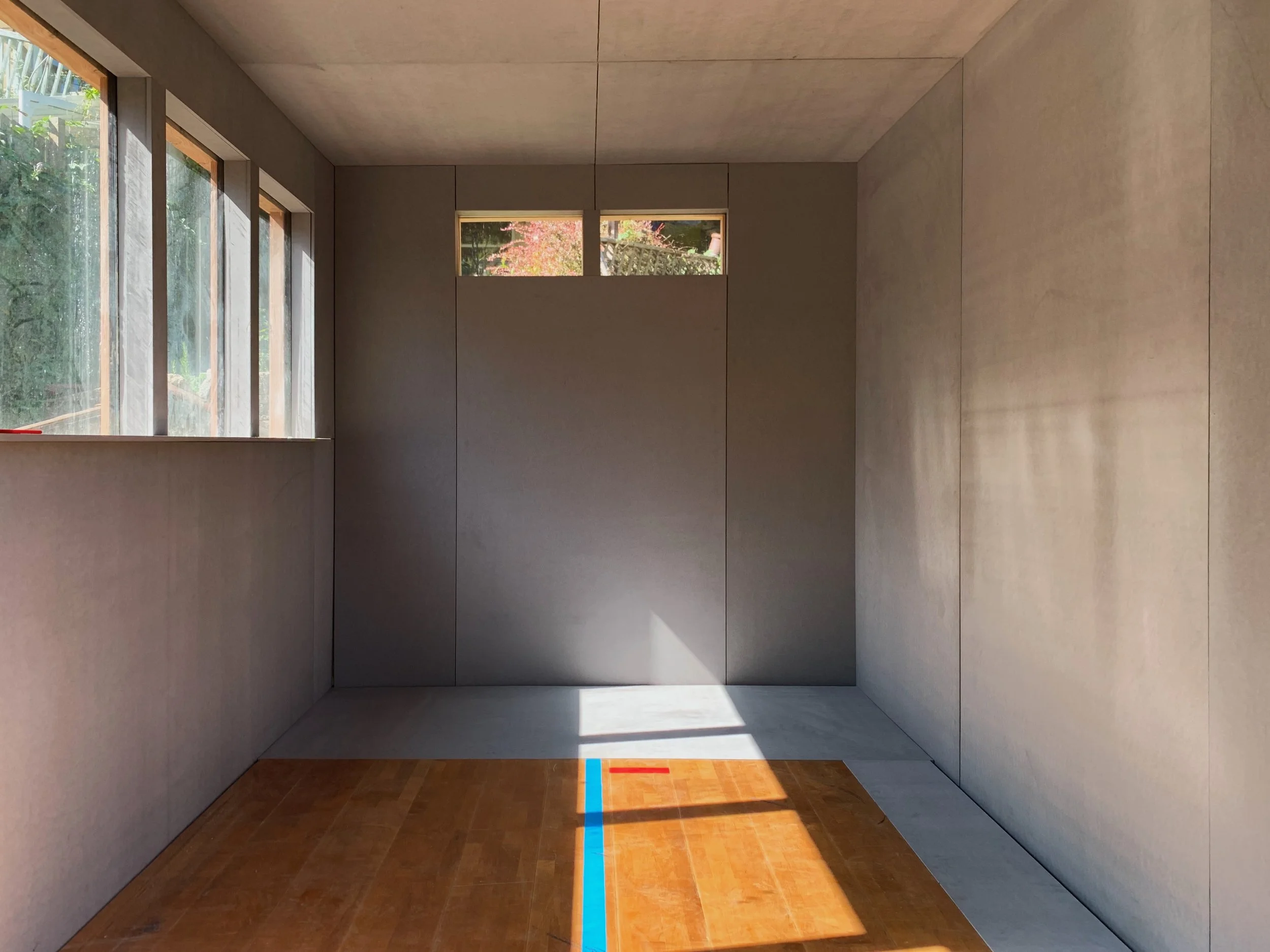 An empty room with light-colored walls, wooden floor, multiple windows on the left side and a small horizontal window near the ceiling at the back, sunlight streaming into the room.