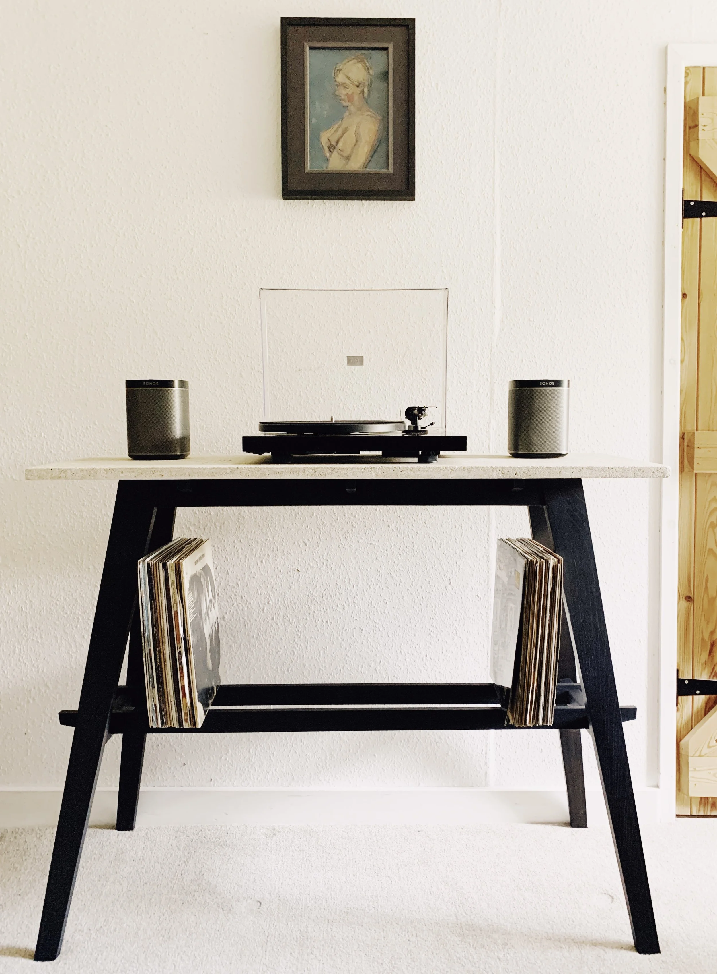 A minimalist turntable with speakers on a black stand against a white wall, with a record shelf below, picture frame on wall.