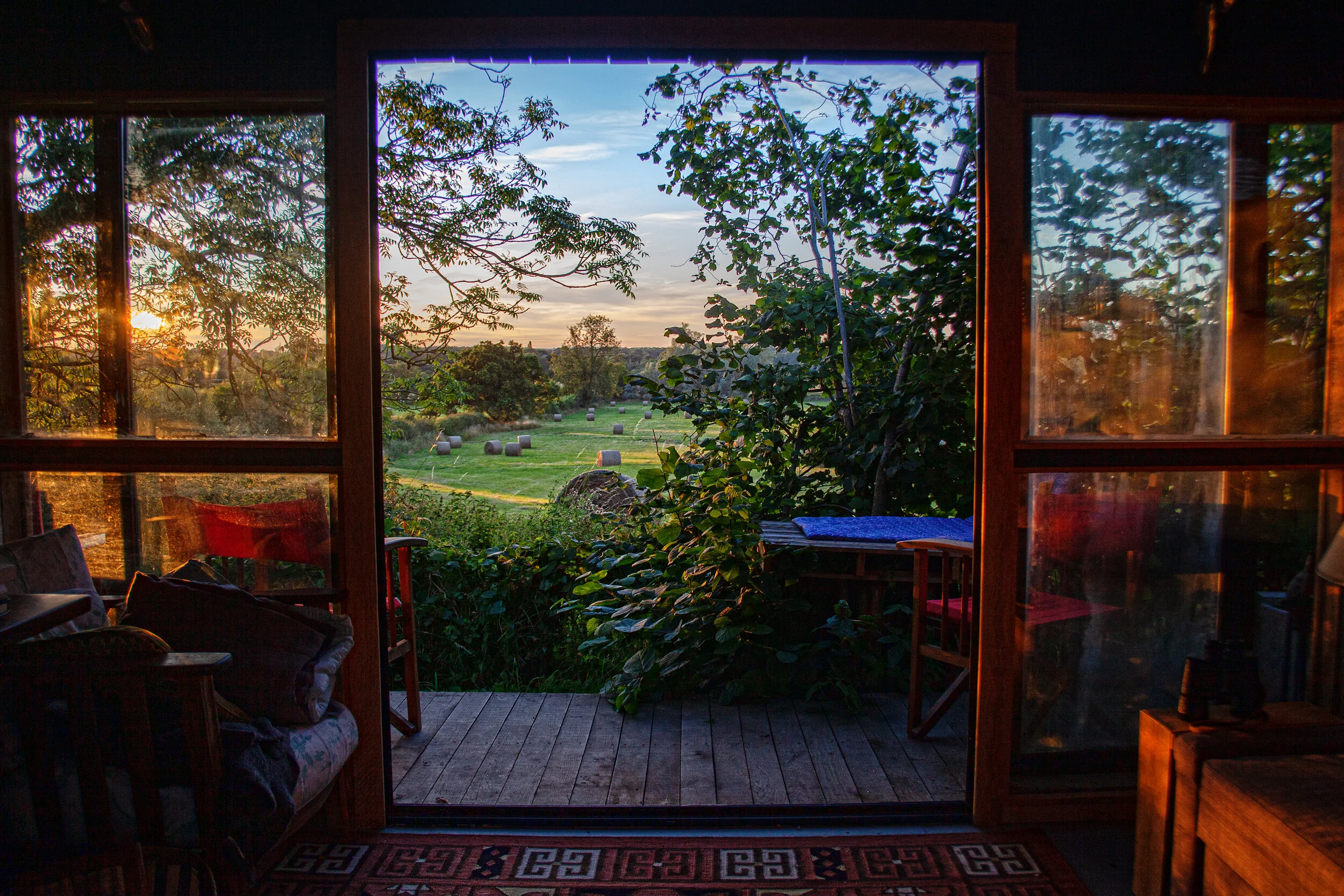 View from inside a house looking out through a glass door at a countryside landscape with a field, hay bales, trees, and a sunset sky.
