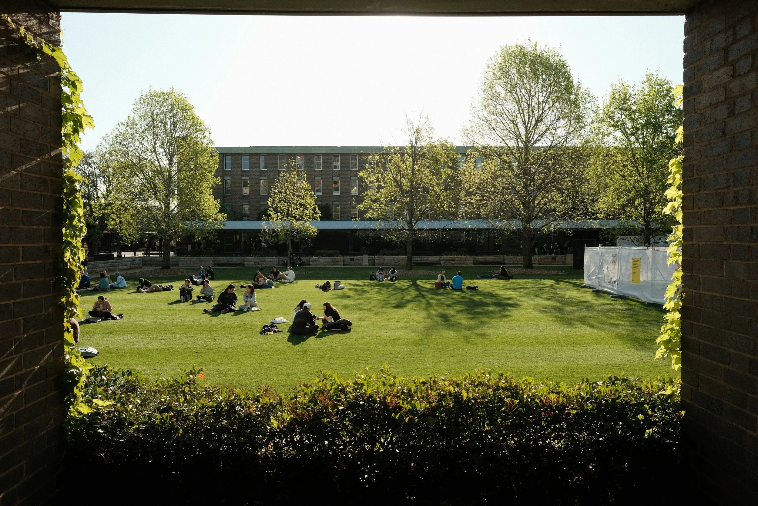 A view through an arch of small groups of students sitting on a lawn at a university campus.