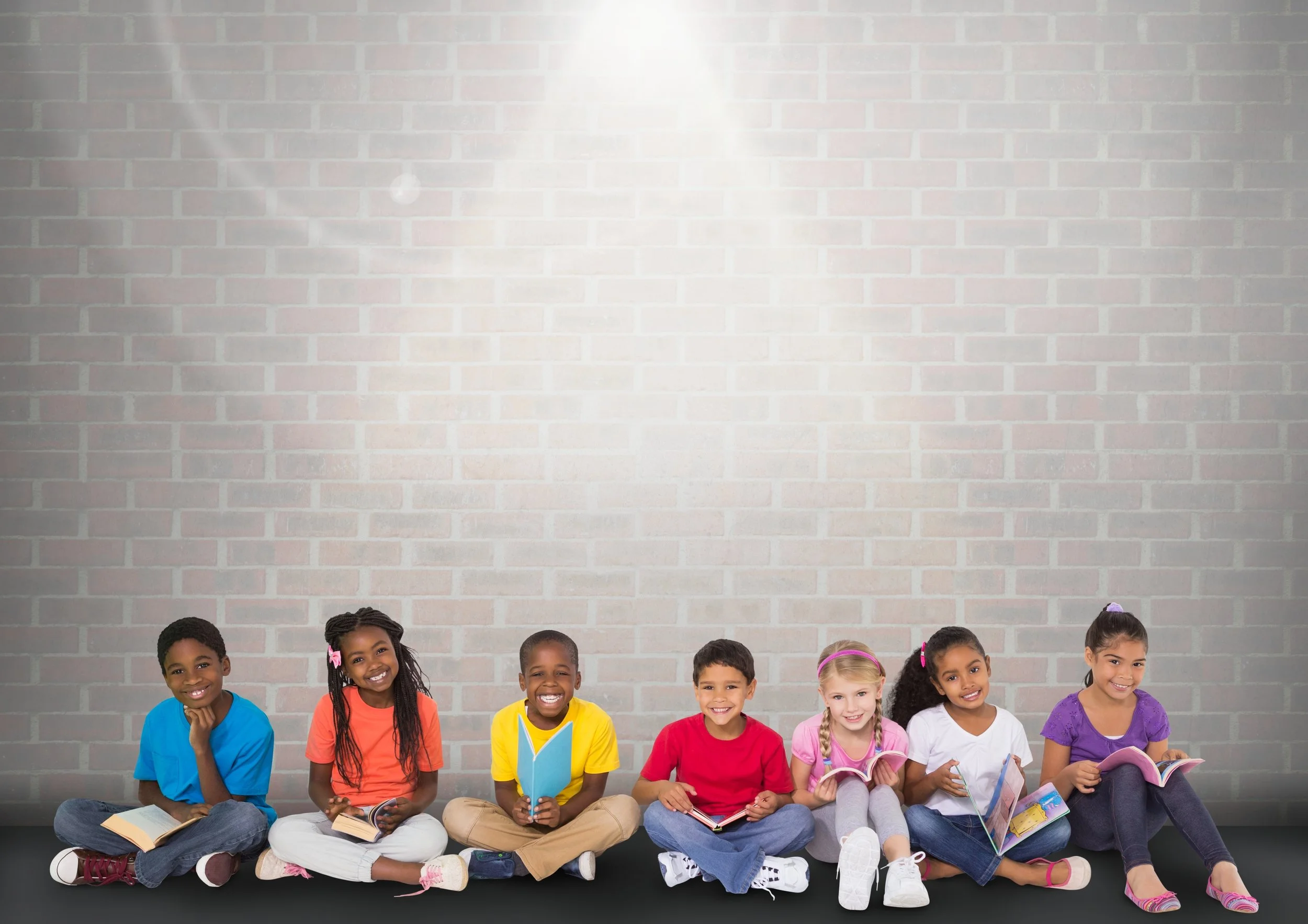 School children with diverse ethnicities are sitting in front of a white wall reading books.