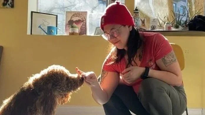 A woman wearing a red beanie and glasses squatting down and playing with a curly-haired dog, in a room with yellow walls and a windowsill with pictures and plants.