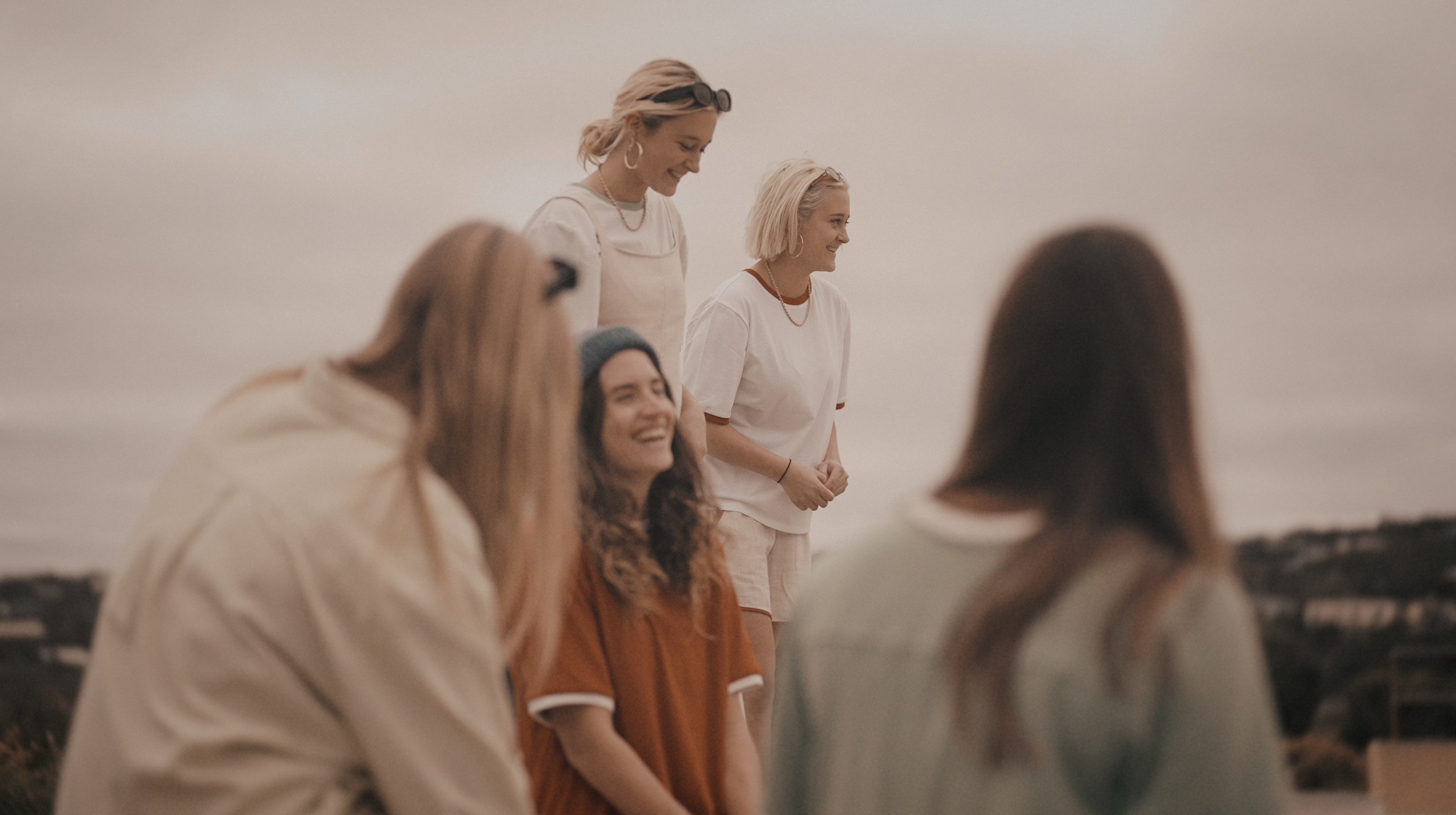 A group of women standing and sitting outdoors at sunset, smiling and chatting.
