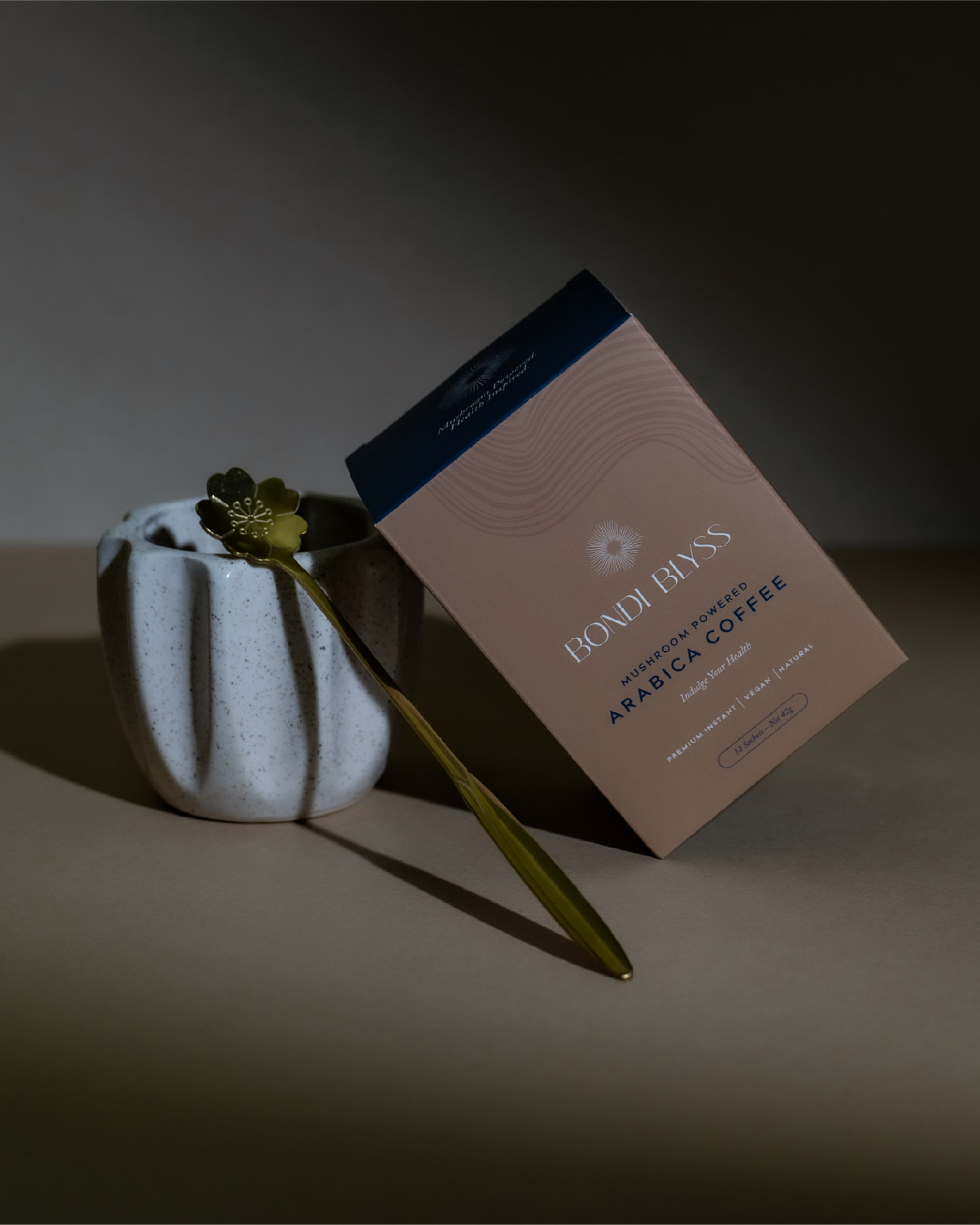 A box of Bondi Bliss mushroom powered Arabica coffee tilted next to a white speckled ceramic pot with a gold flower-shaped spoon resting across it, all on a beige surface with shadowed background.