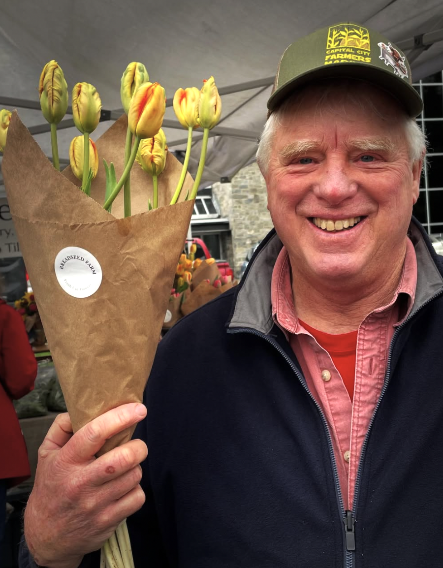 A smiling man holding a bouquet of yellow and orange tulips wrapped in brown paper at an outdoor market.