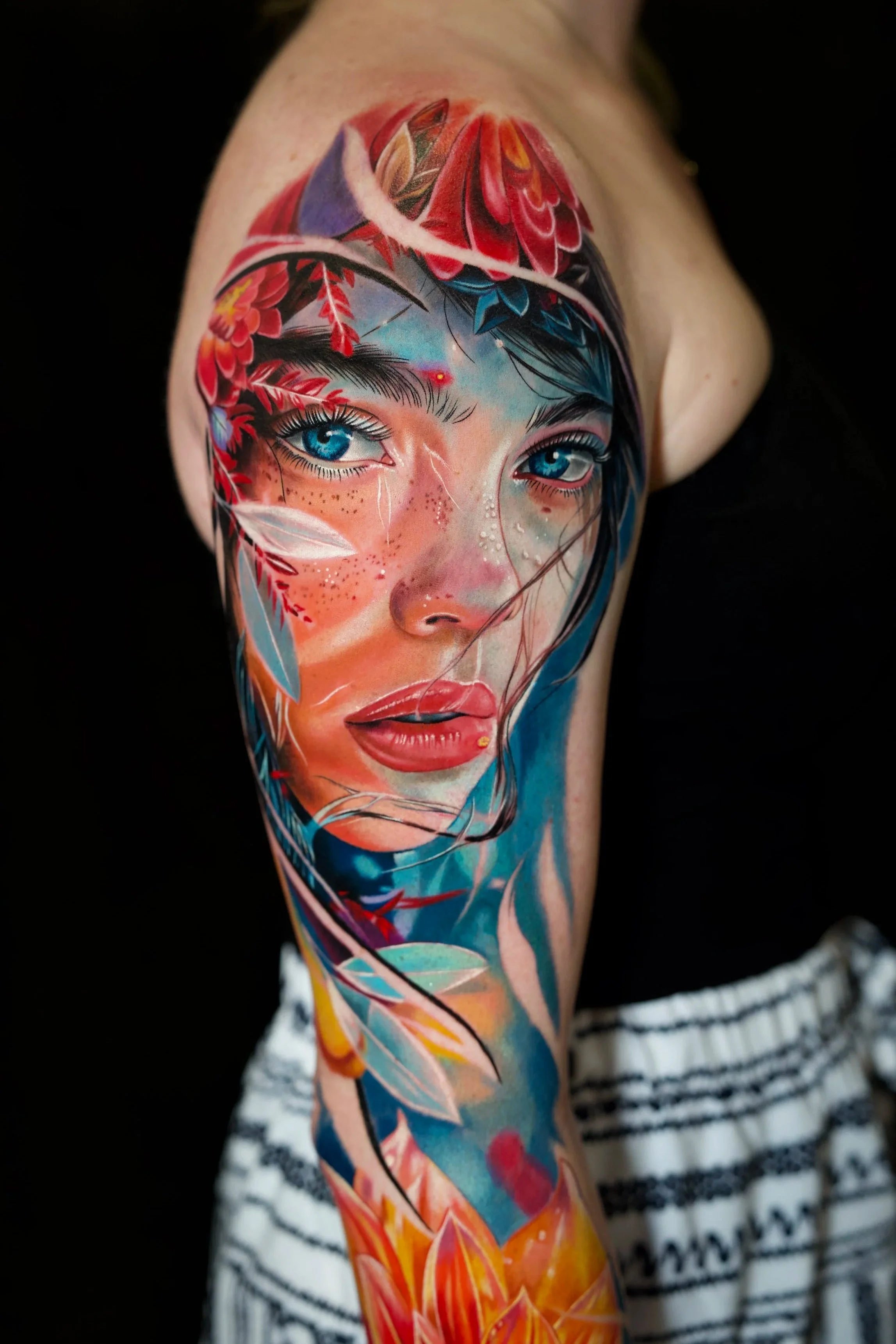 Colorful tattoo of a woman's face with blue eyes, surrounded by vibrant leaves and floral elements, covering the upper arm and shoulder.