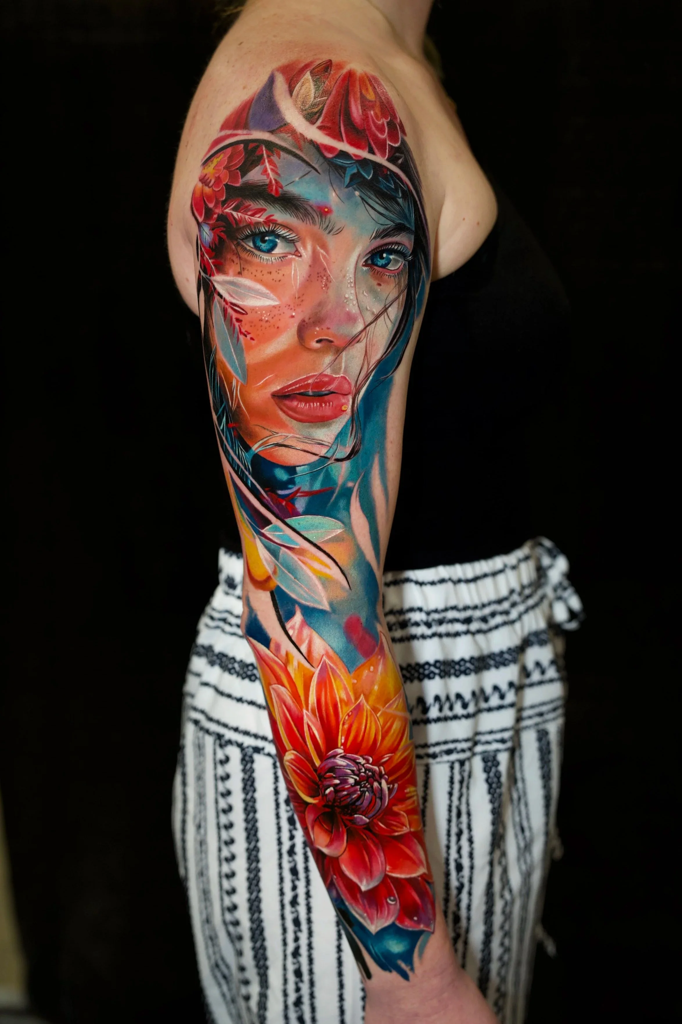 Colorful tattoo of a woman's face with blue eyes, surrounded by flowers, leaves, and abstract elements on the person's arm.