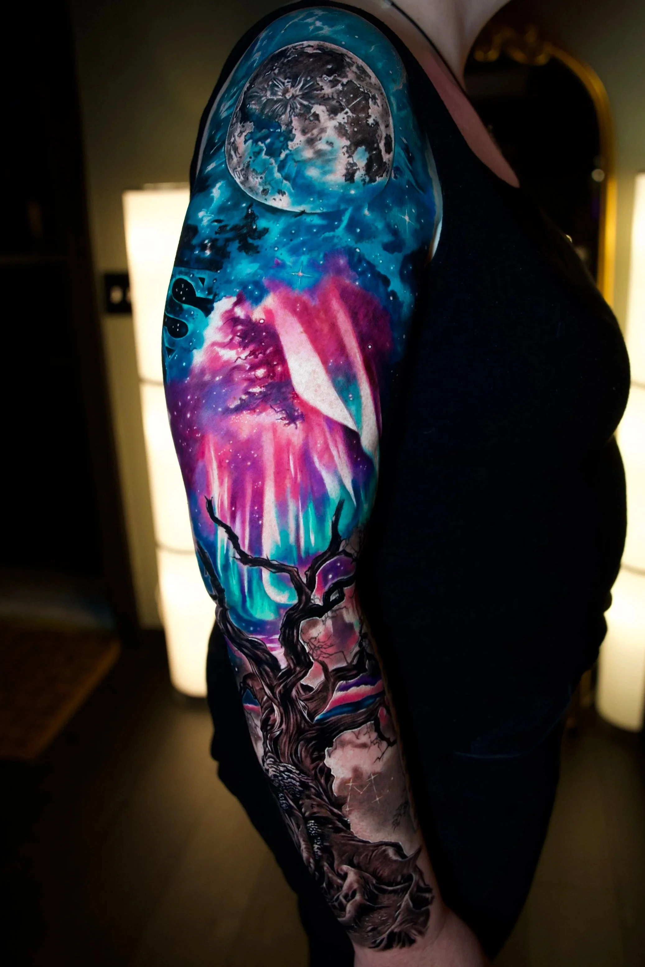 Detailed colorful tattoo sleeve on a person's arm, depicting a cosmic scene with a full moon, stars, aurora lights, and a twisted tree.