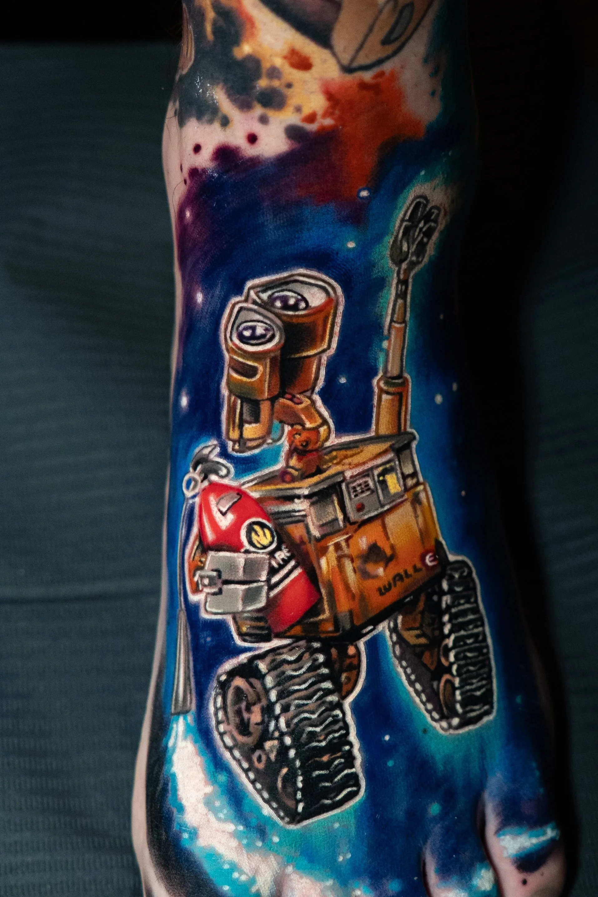 A colorful tattoo of WALL-E, the animated robot, on a person's arm, set against a cosmic background.