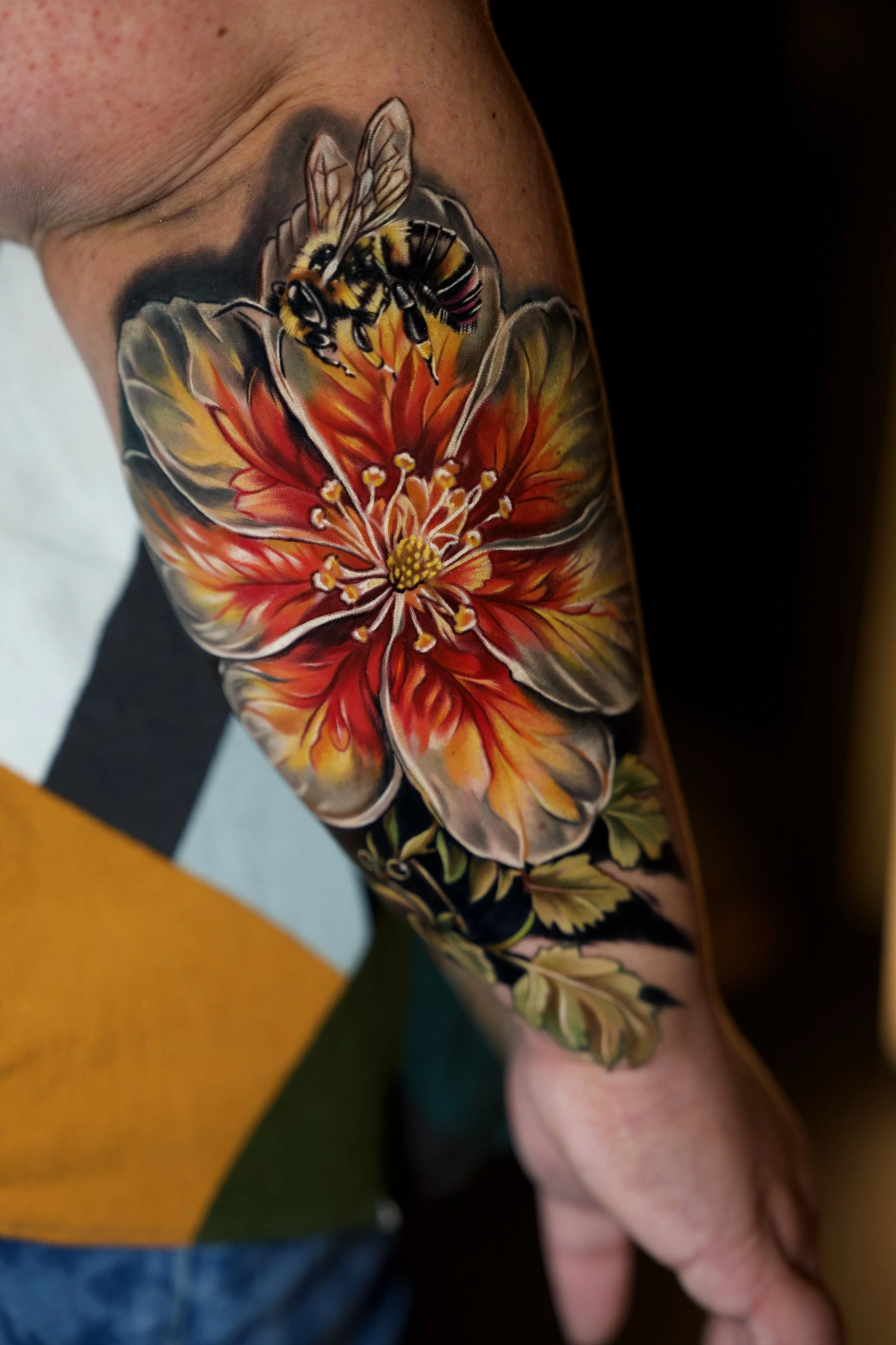 Close-up of a detailed tattoo on a person's arm featuring a bee hovering over a large orange and pink flower with green leaves.