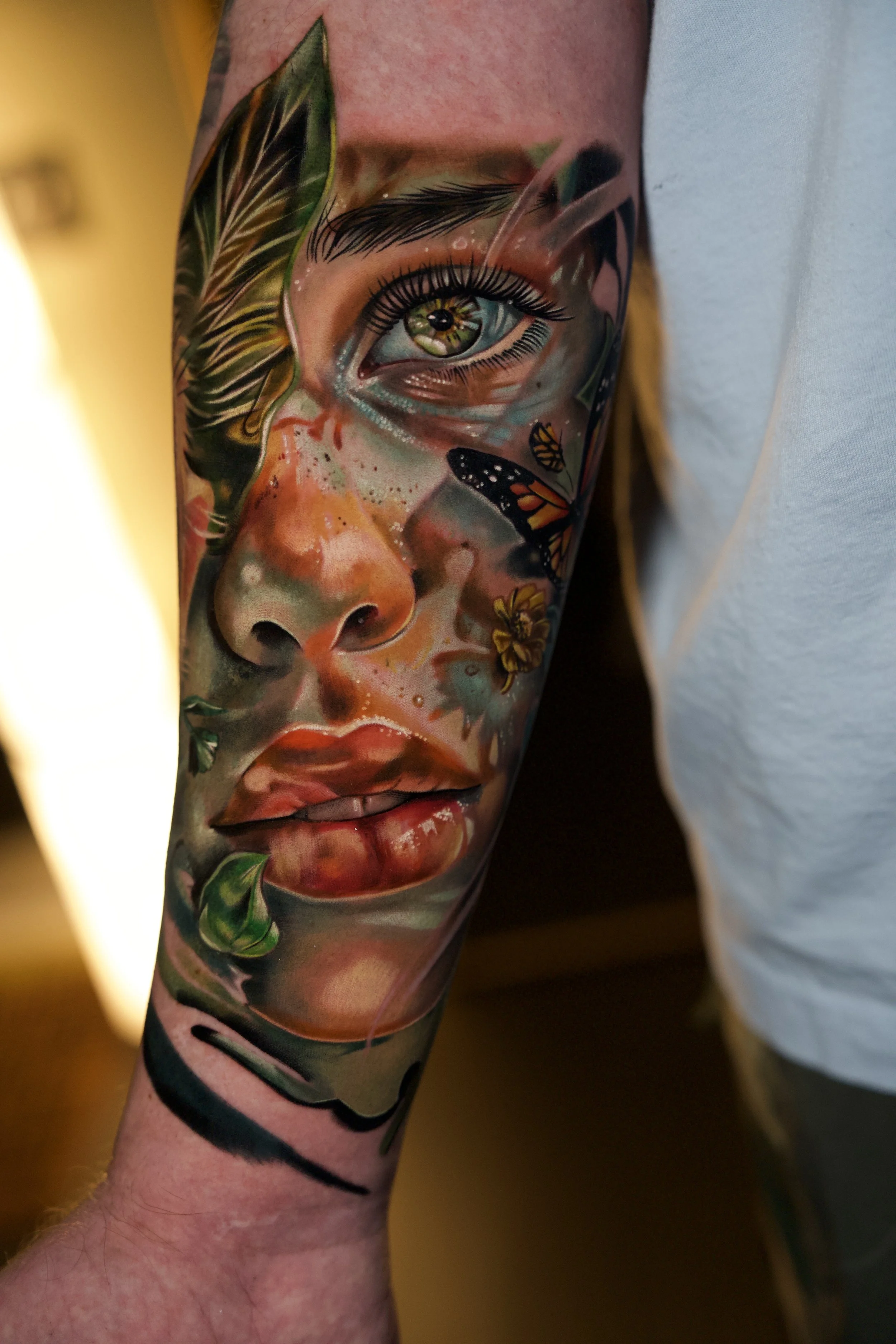 Close-up of a colorful realistic tattoo of a woman's face on a person's arm, with detailed features including green eyes, full lips, and decorative elements like butterflies, leaves, and vibrant colors.
