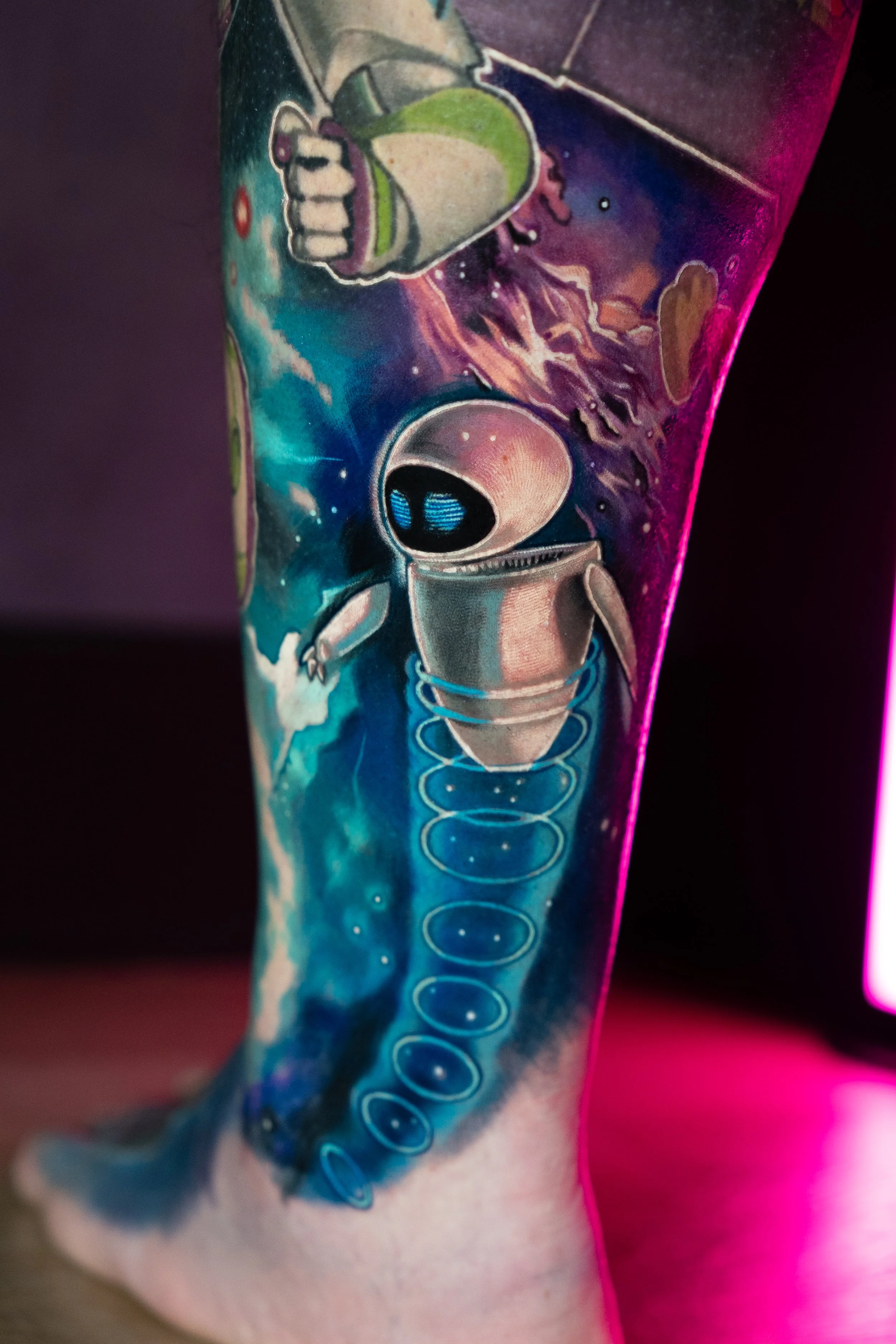 A colorful tattoo on a person's leg featuring Eve from Wall-E. The background shows a cosmic scene with planets, stars, and nebulae.