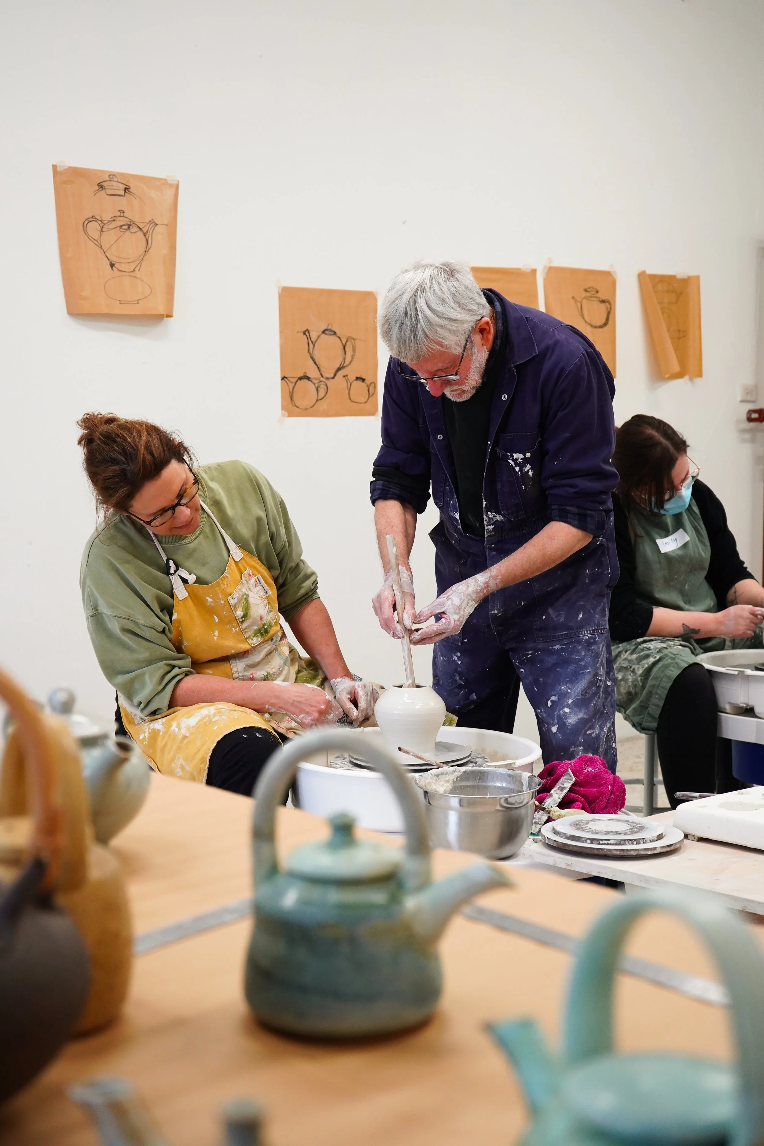 People participating in a pottery class, working on pottery pieces at a table in an art studio, with sketches of teapots and cups on the wall behind them.