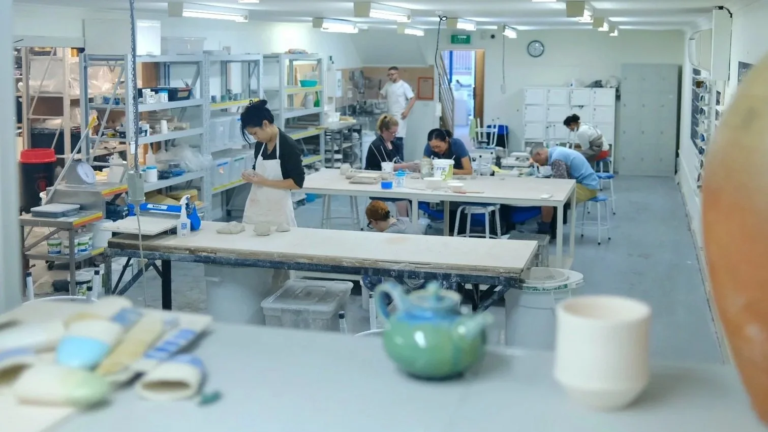 People working in a ceramics or pottery workshop, shaping clay and working at tables with various tools and supplies, in a well-lit studio.