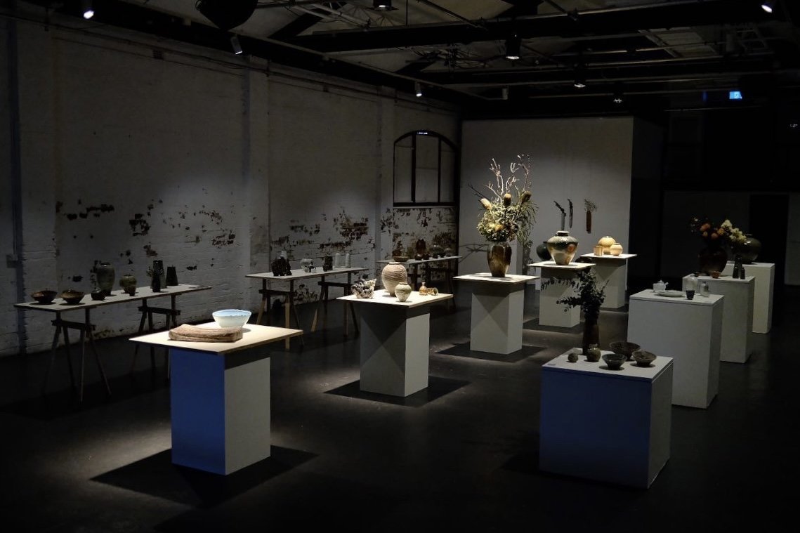An art gallery featuring various ceramic vases, bowls, and sculptures displayed on white pedestals and tables in a dimly lit room.