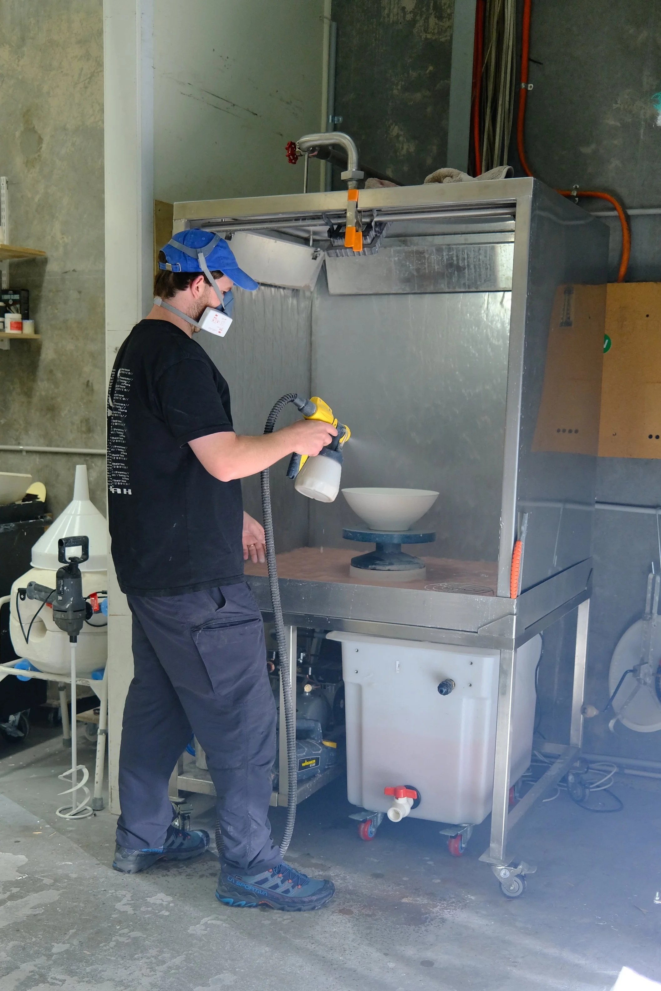 A person wearing safety glasses, a mask, and a blue cap is using a spray gun in a ceramic kiln. The kiln is a metal industrial-type with a ceramic bowl inside, surrounded by a protective cover, in a workshop setting.