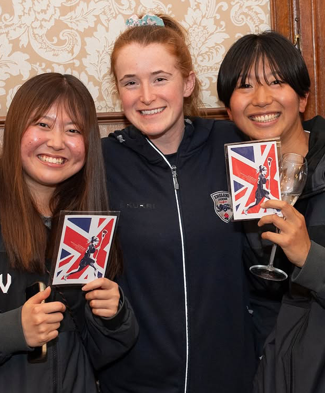 Three lacrosse players — one from England and two from Japan — holding the mini postcard version of the England Lacrosse Home Internationals poster.