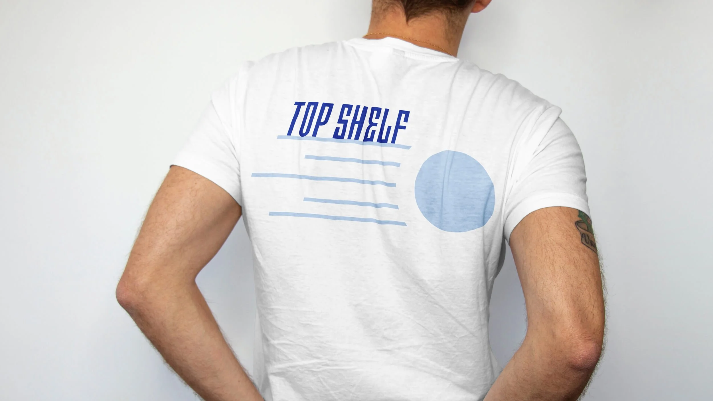 A person from the back wears a white t-shirt with "TOP SHELF" printed in blue on the upper back, along with blue horizontal lines and a blue circle.