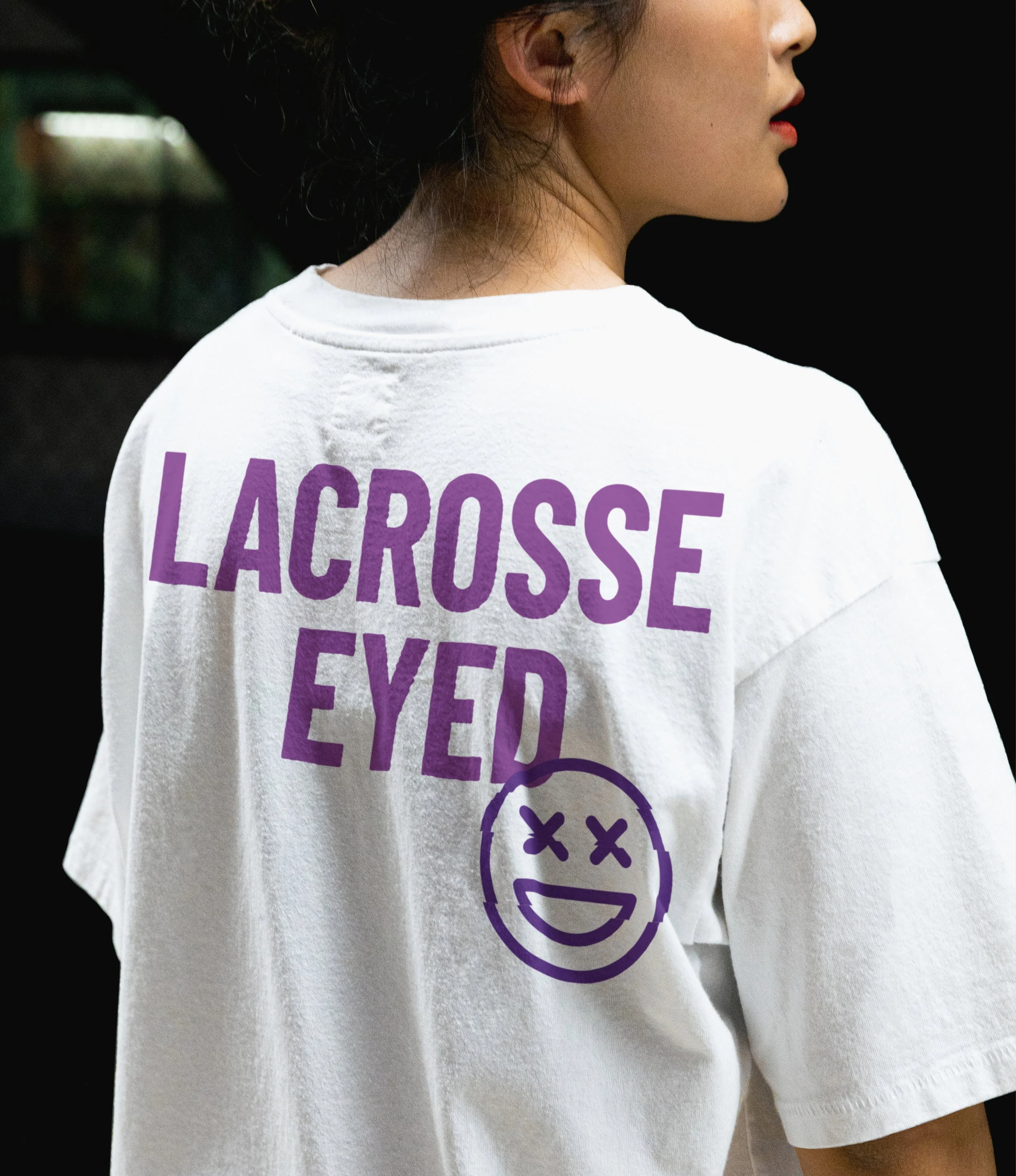 A person wearing a white t-shirt with the purple text "LACROSSE EYED" and a graphic of a smiley face with "XX" eyes.