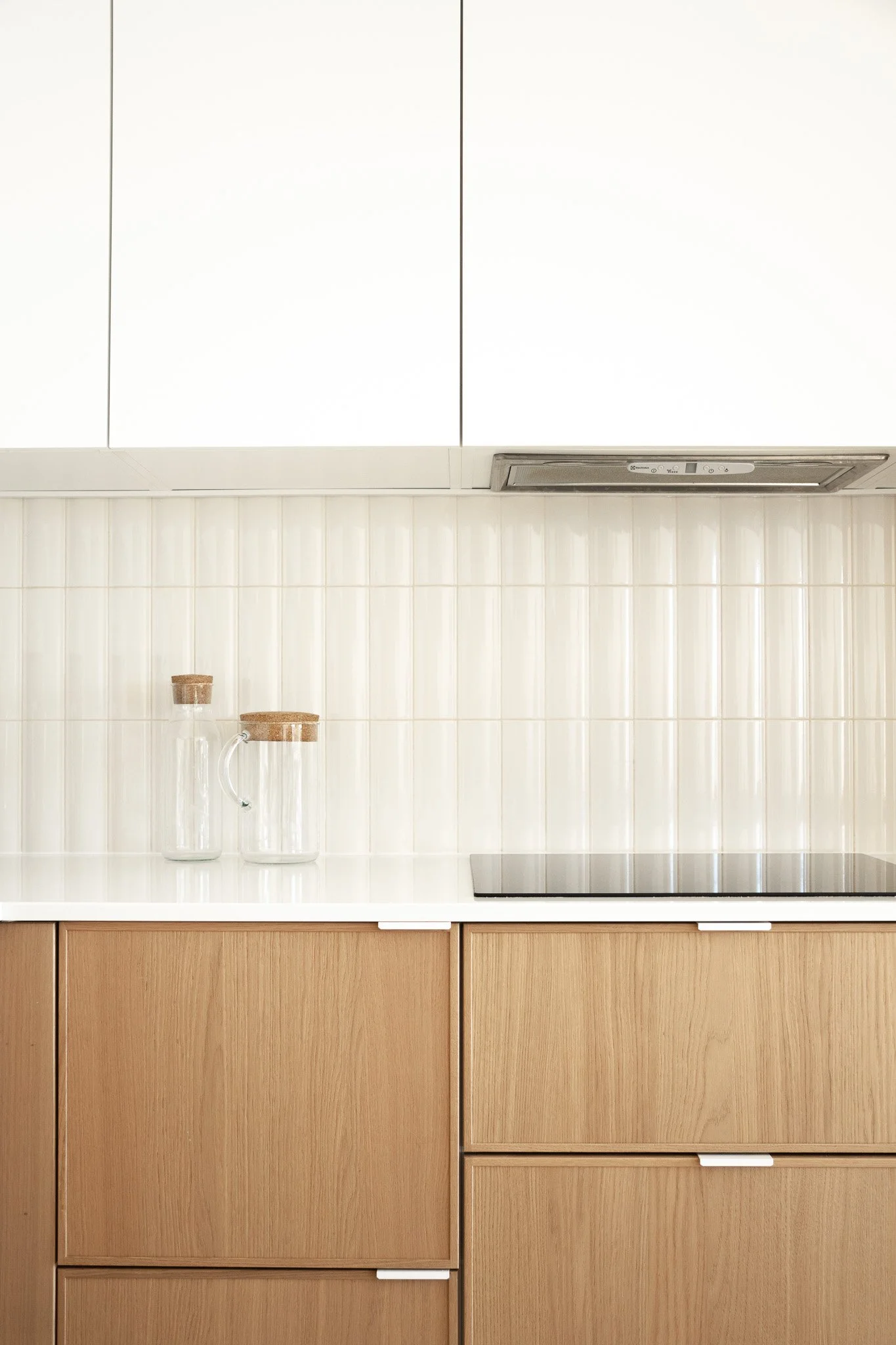 Cuisine minimaliste avec armoires en bois clair, comptoir blanc, deux jarres en verre avec couvercles en bois, plaques de cuisson noires, et un mur en carrelage beige éclatant.