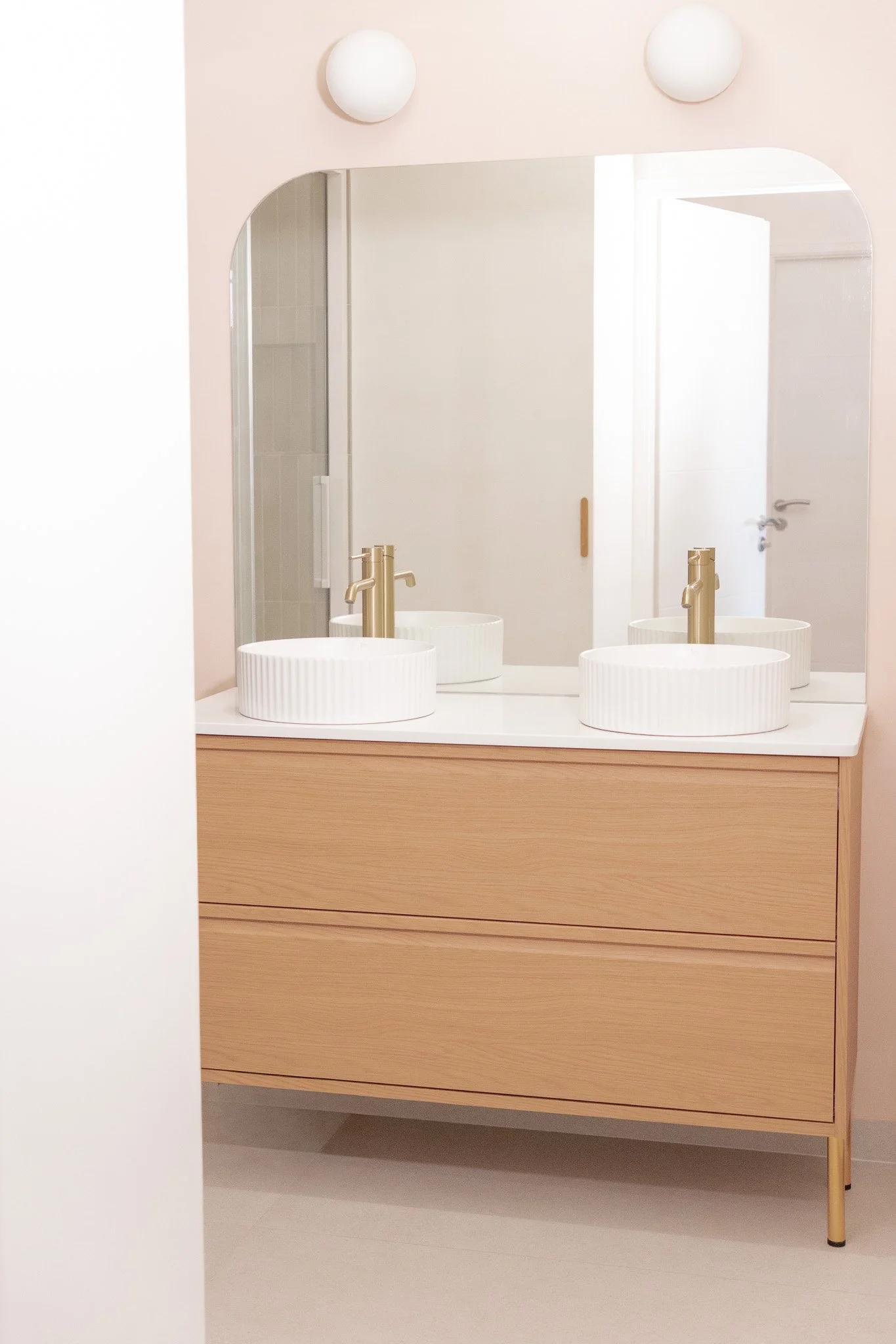 meuble de salle de bain en bois clair avec deux tiroirs, deux lavabos ronds en céramique blanche à robinetterie or antique, miroir mural au-dessus, et murs rose poudré.