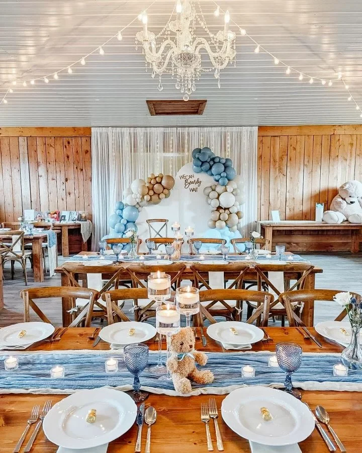 Decorated indoor event space with a wooden wall, white curtains, and a chandelier, featuring a balloon arch in blue, white, and beige, and a table setup with plates, glasses, and candles.