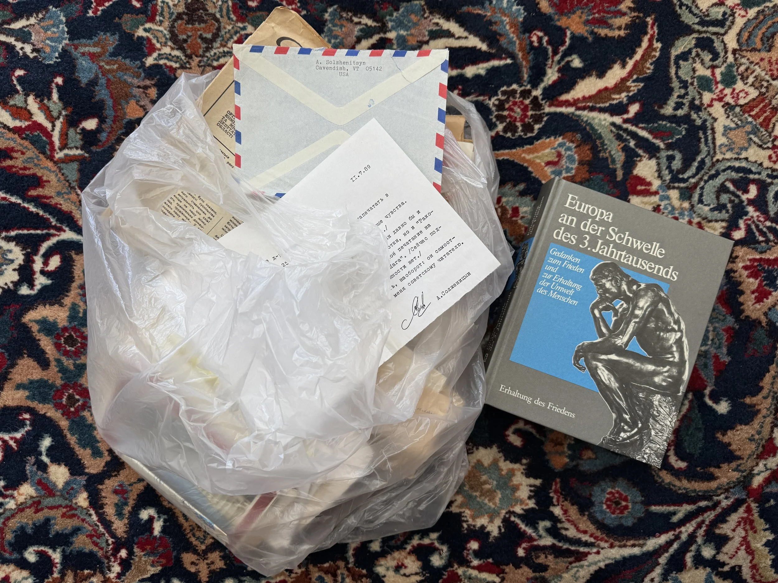 A plastic bag filled with old family letters lies on a patterned carpet next to the book “Europa an der Schwelle des 3. Jahrtausends.” One visible envelope is addressed from Alexander  Solzhenitsyn