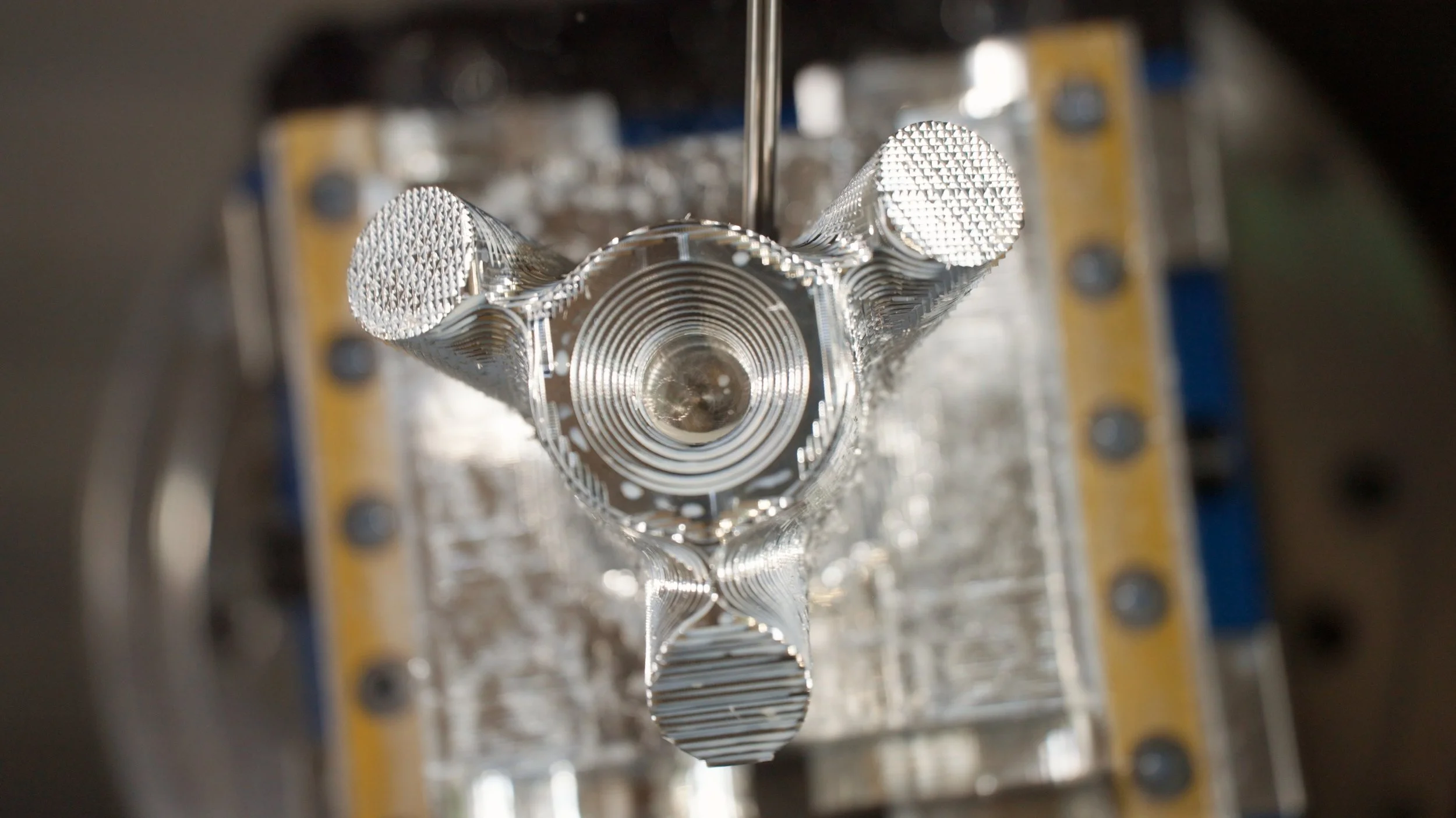 A metallic component with a circular pattern in the center, surrounded by three cylindrical structures, mounted on a base with bolts.